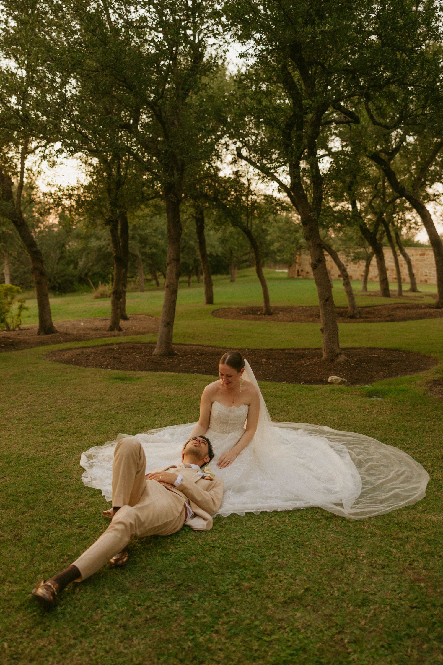 Bride in white gown and groom in tan suit relaxing on green grass surrounded by trees at golden hour.