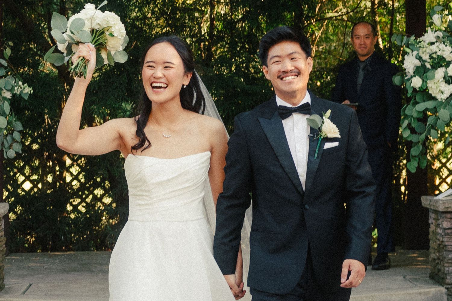 Happy Asian couple in wedding attire, bride in white gown holding bouquet, groom in tuxedo, outdoor ceremony.