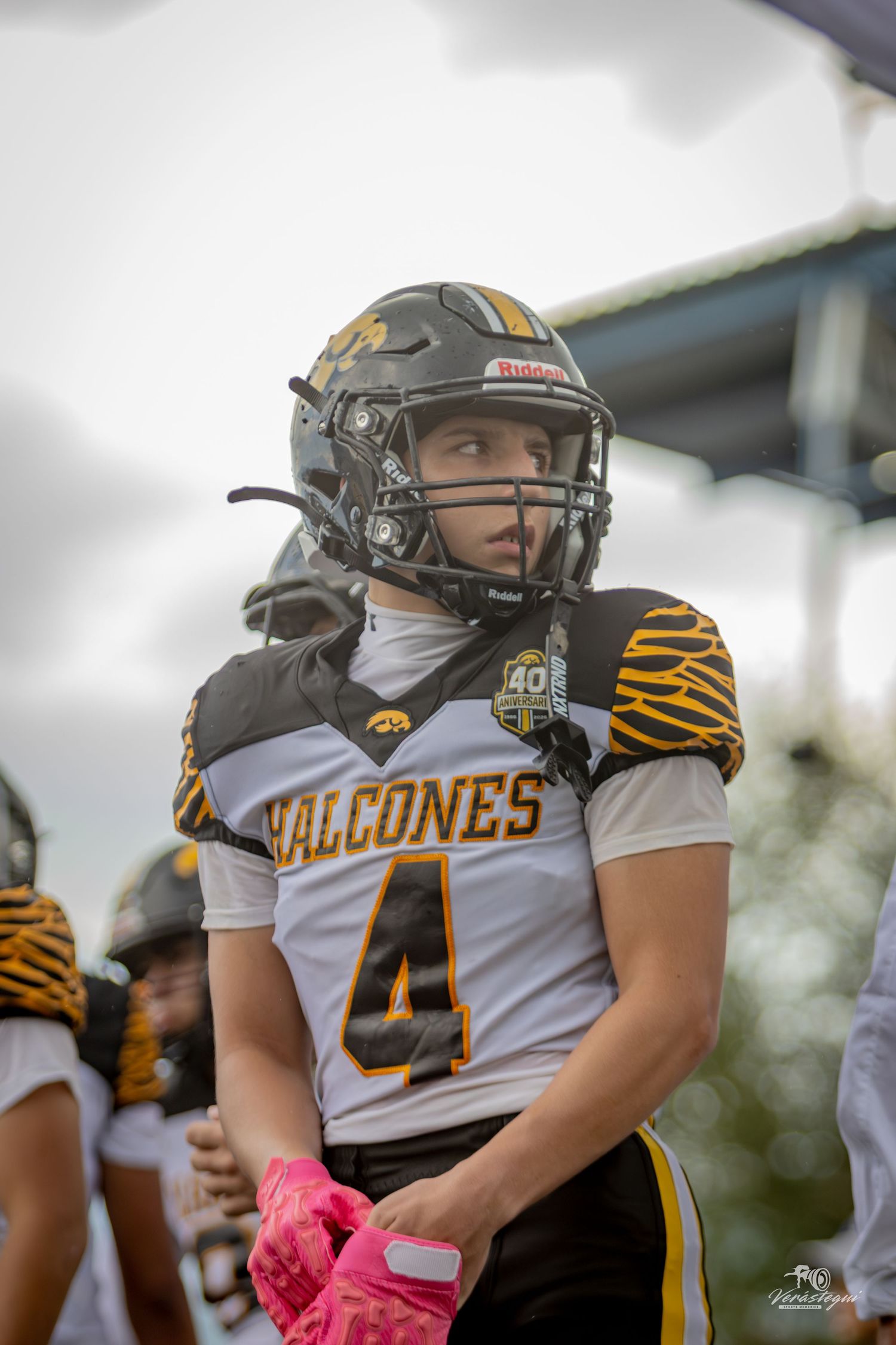 Young football player wearing a Halcones #4 jersey and helmet stands on the sideline during a game.