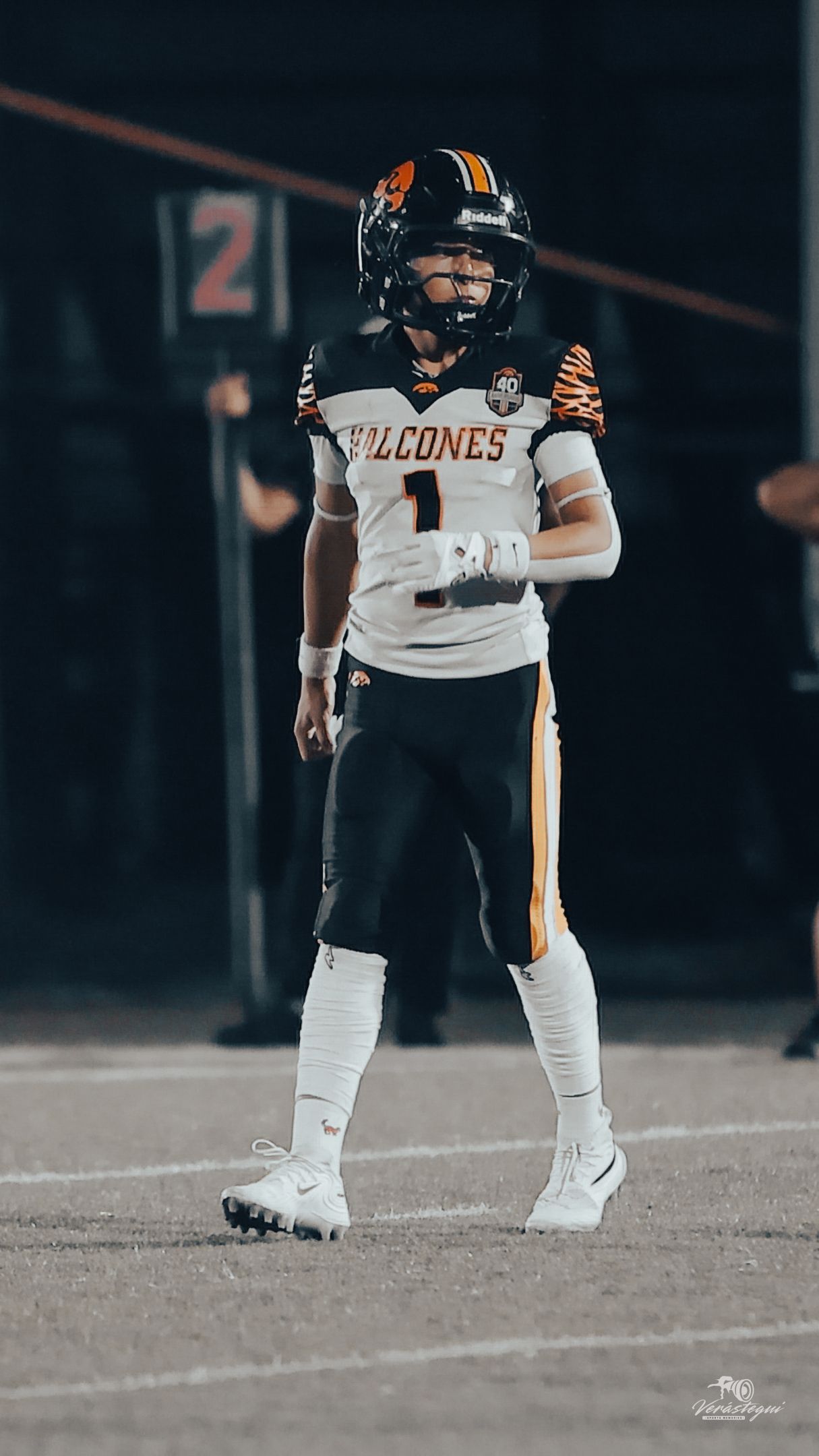Falcones football player wearing jersey number 1 in white and black uniform with orange accents stands on field at night.