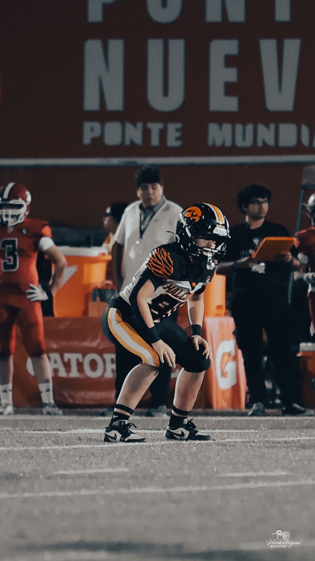 Young football player in black and orange uniform crouches in ready stance on field during night game.