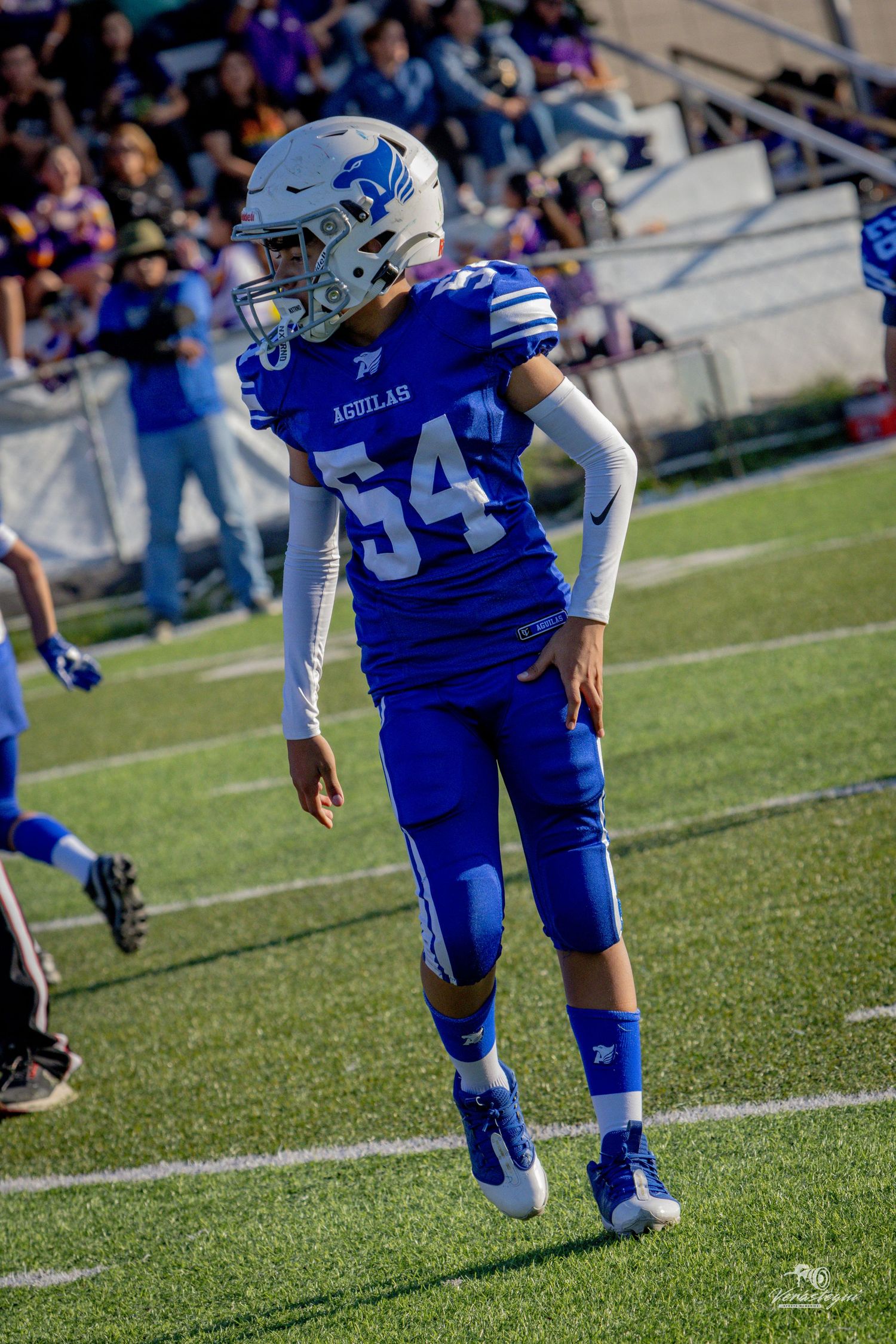 Youth football player #54 in blue Aguilas uniform and helmet stands on field during game.