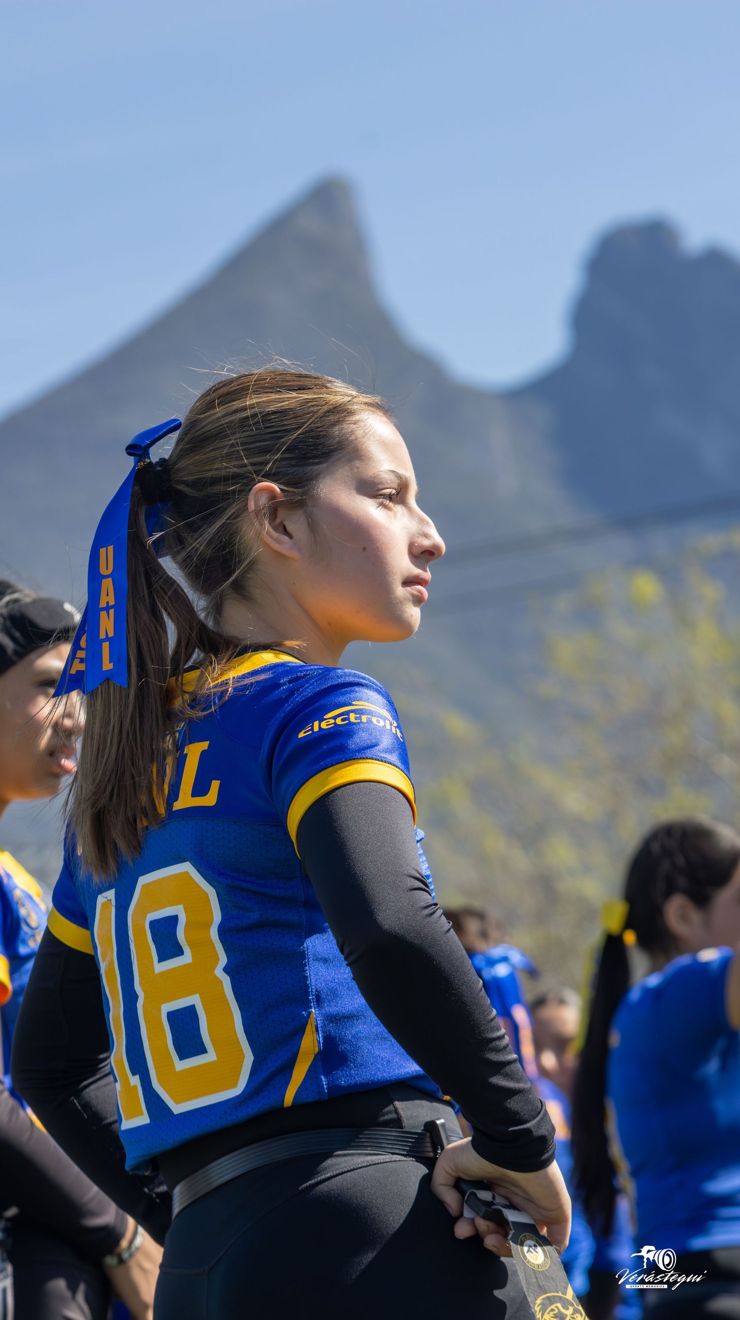 Softball player in blue and gold uniform stands against mountain backdrop during outdoor game.