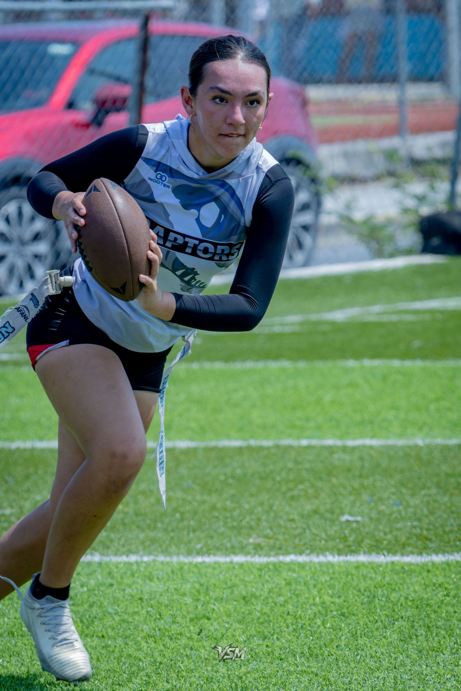 Female flag football player in Raptors jersey runs with ball during outdoor game on turf field.