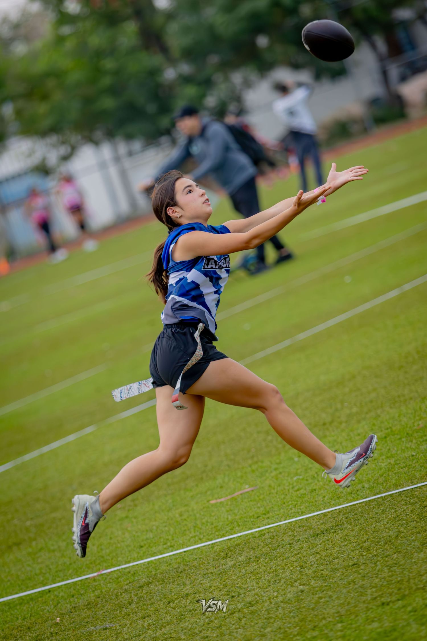 Female athlete leaping to catch a football during an outdoor flag football game on a green field.