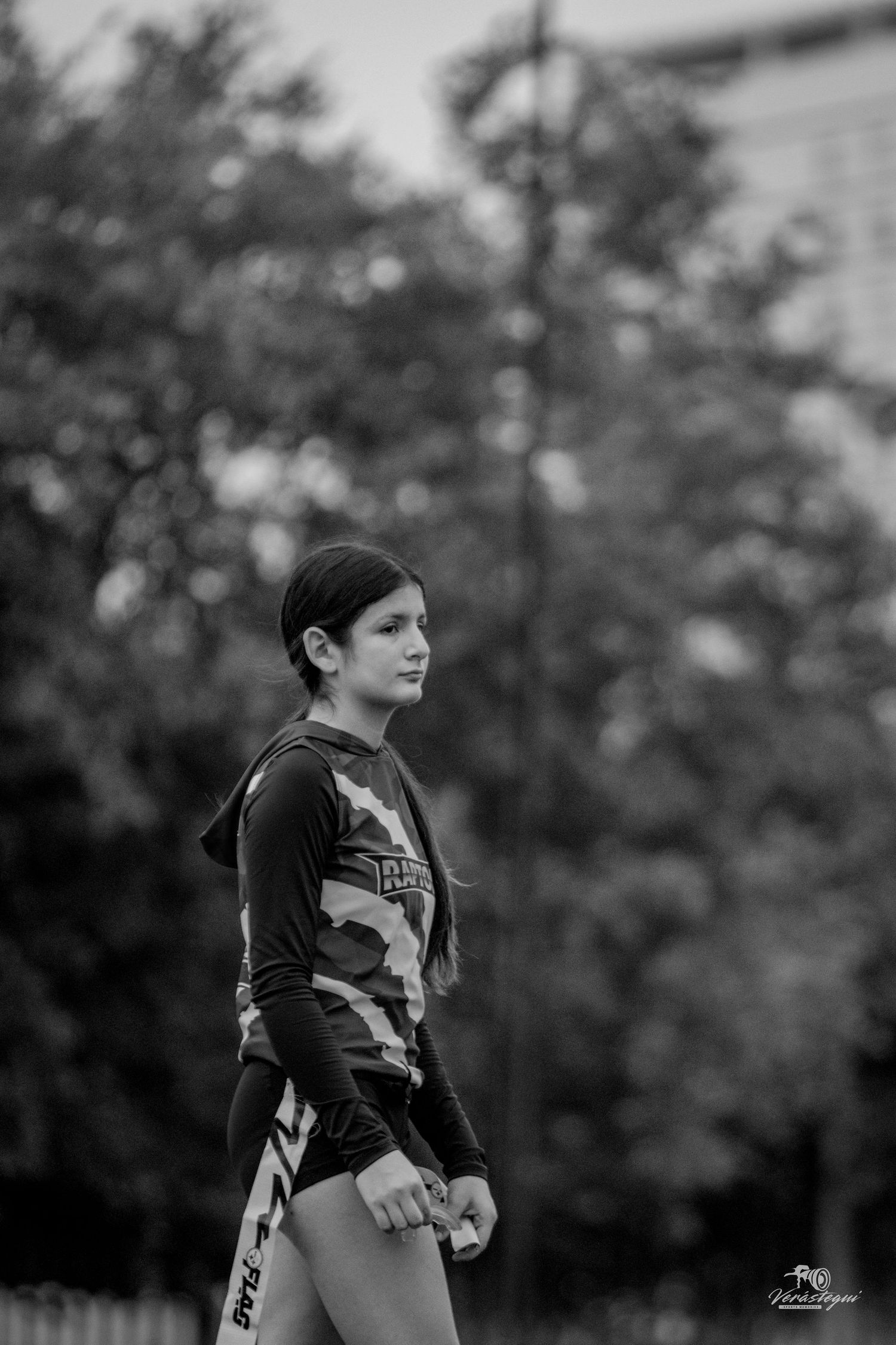Black and white photo of a young athlete in camouflage sportswear standing outdoors with blurred trees in background.