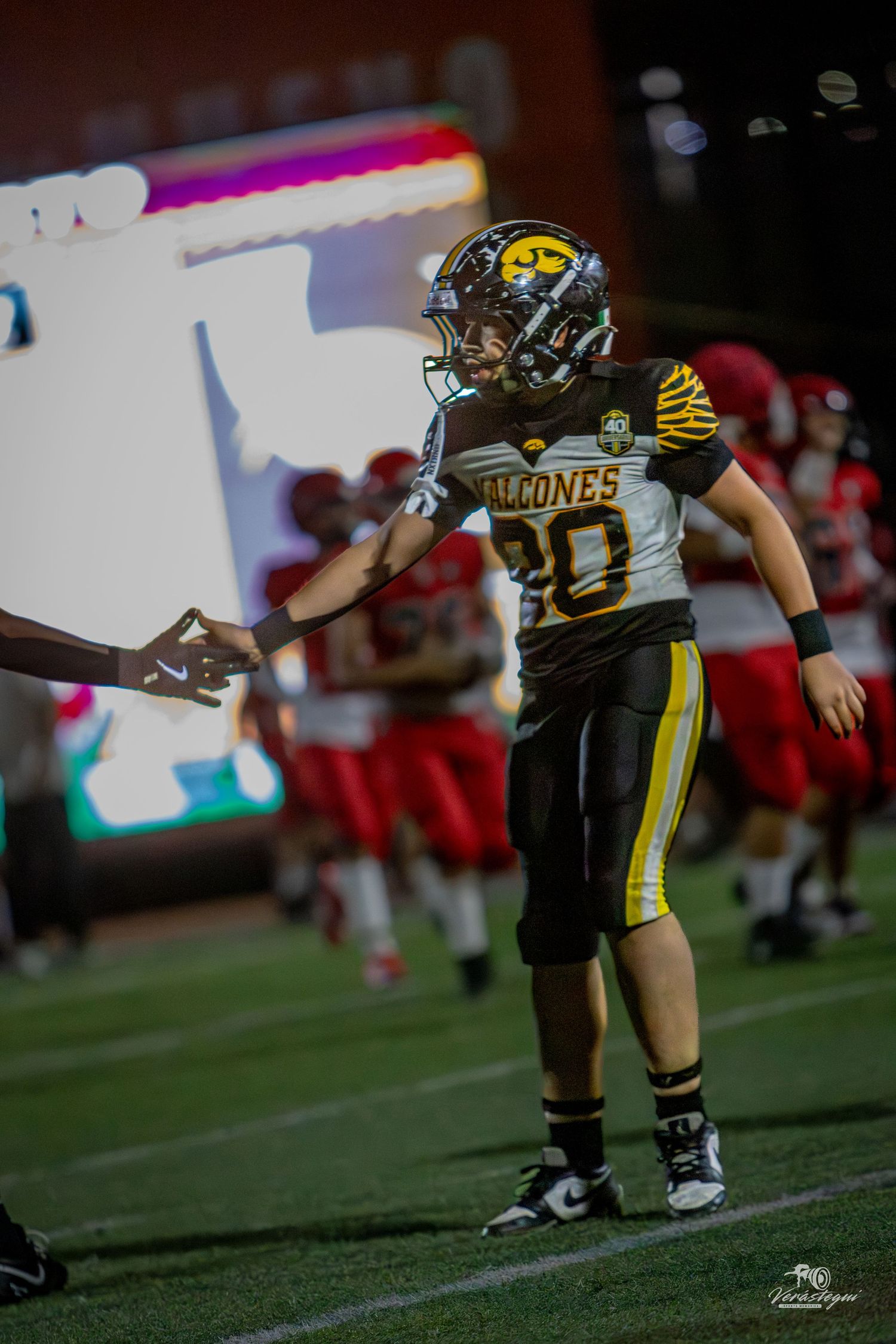 Football player #20 in black and gold Falcons uniform fist-bumping on field during night game.