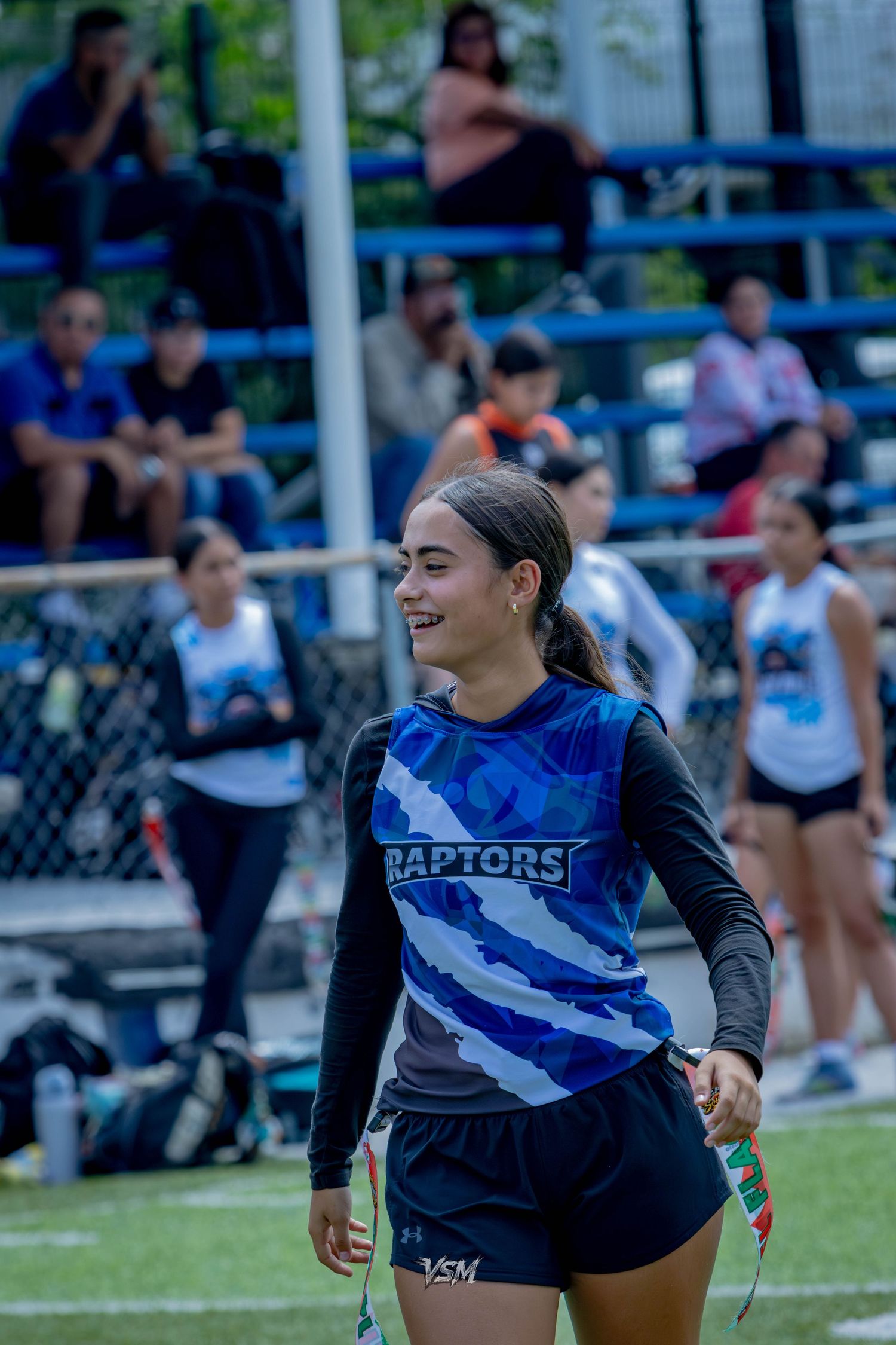 Smiling young woman in blue Raptors jersey playing flag football on outdoor field with spectators in bleachers behind her.