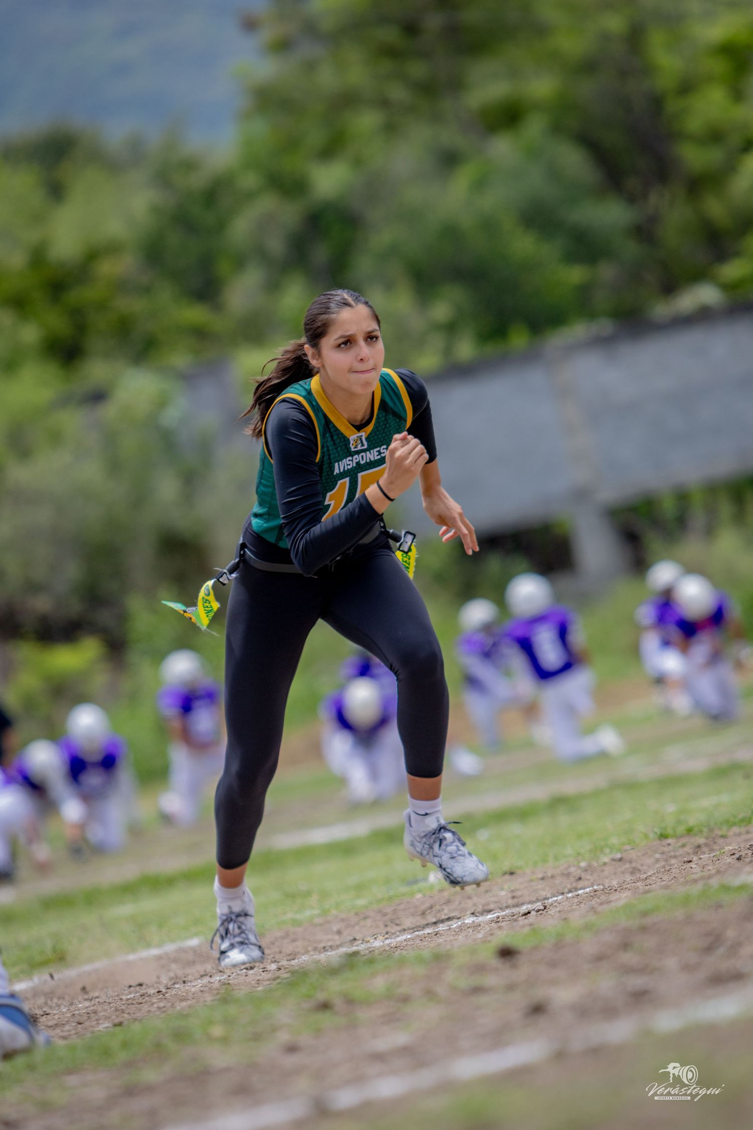 Female flag football player in green jersey sprinting on field during game with purple-uniformed players in background.