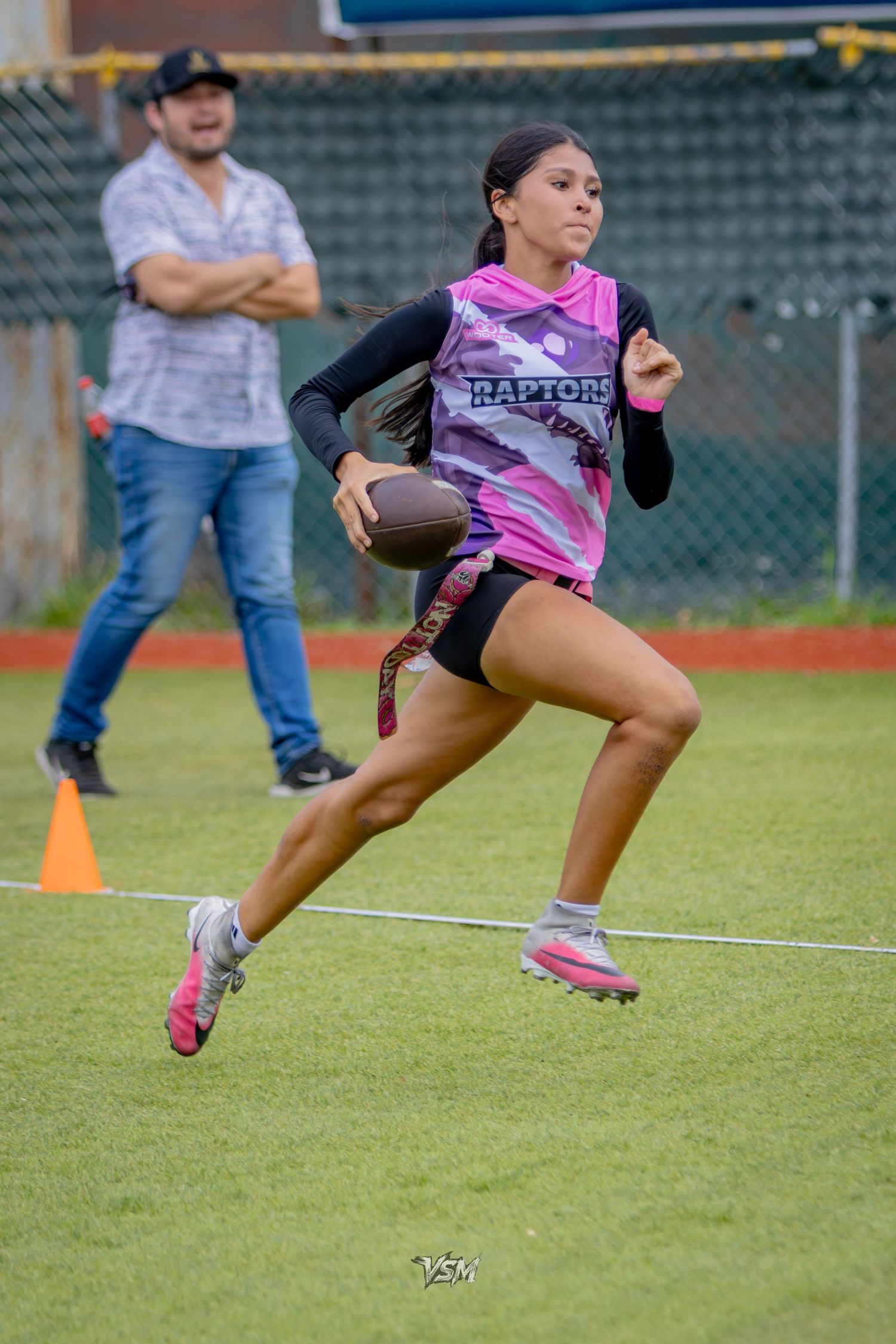 A girl in a pink Raptors jersey runs with a football during a flag football game on a green turf field.