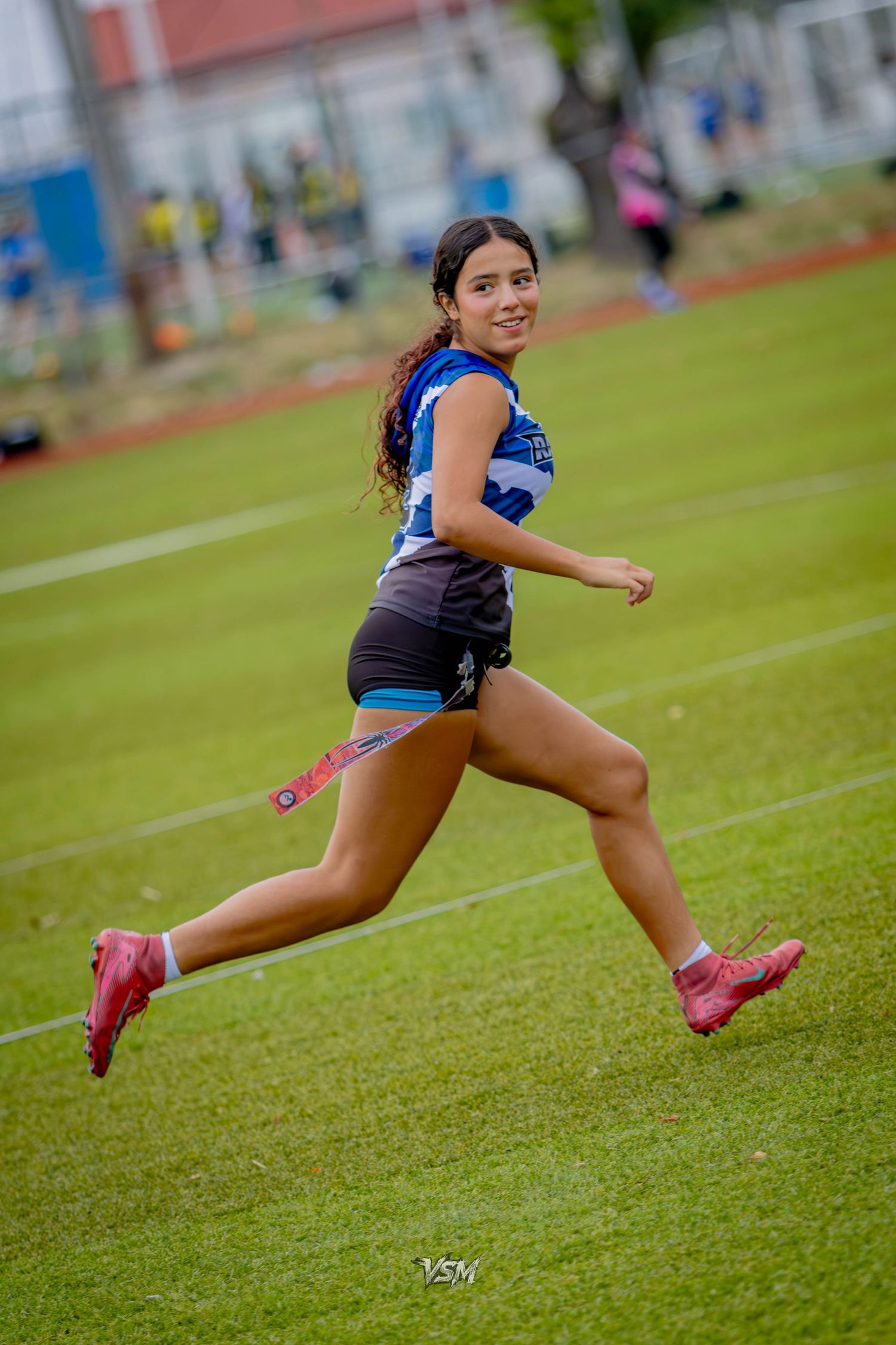 Young female athlete running on green field in blue jersey, black shorts, and red cleats during outdoor sports event.