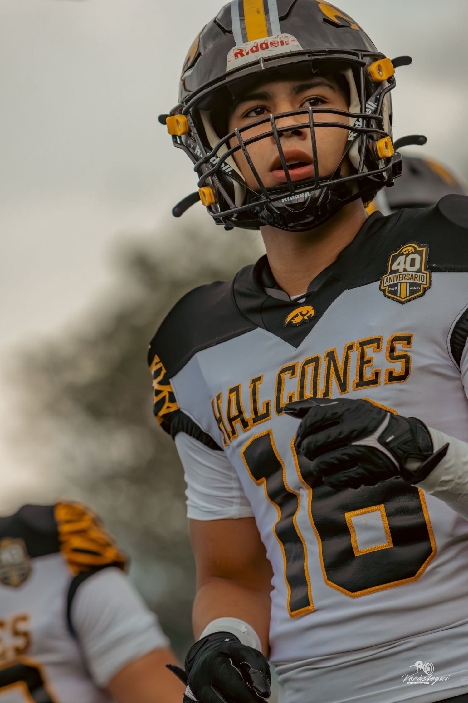 Football player #18 in Halcones uniform and Riddell helmet stands ready on the field during a game.