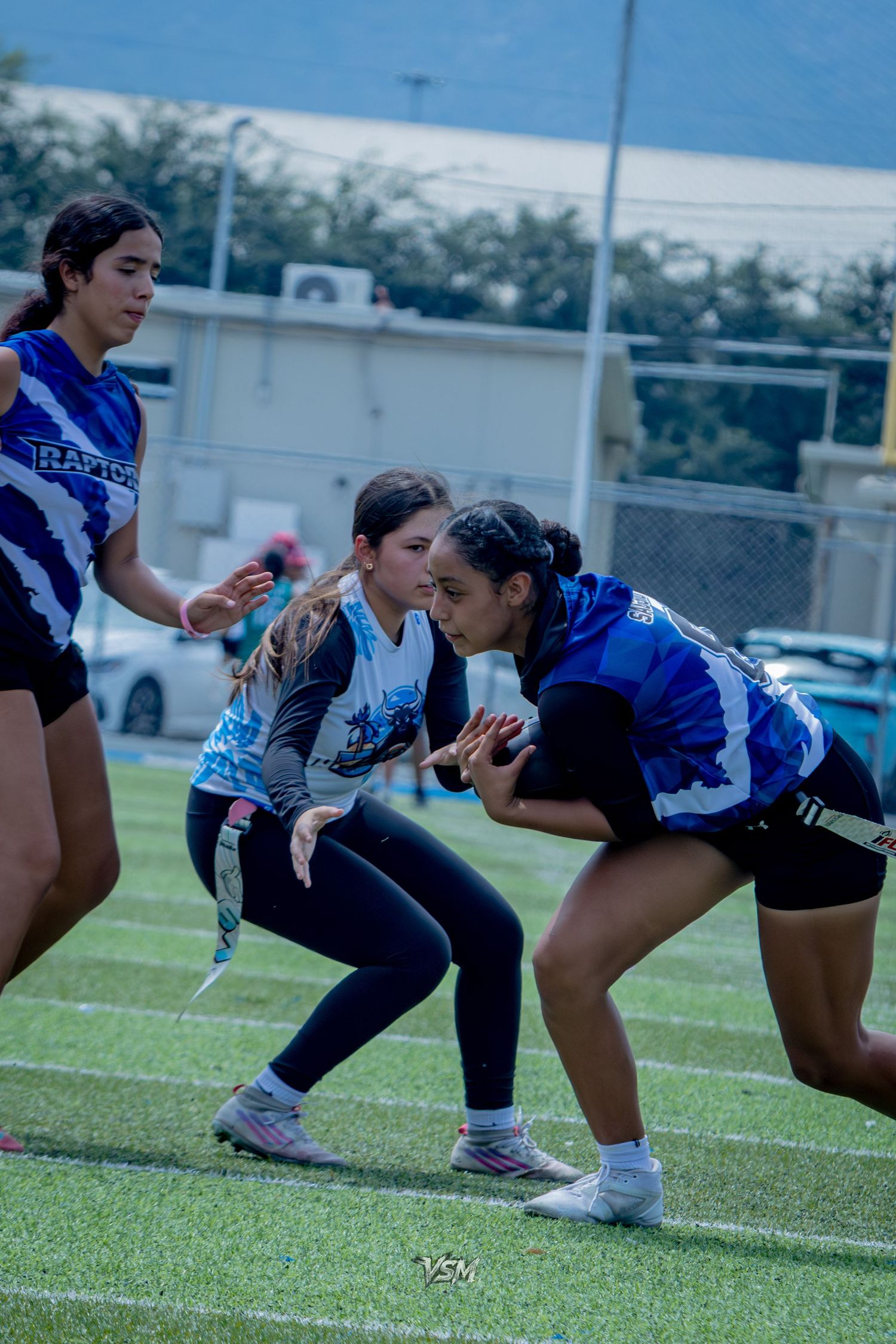 Women playing flag football on a turf field, with players in blue jerseys competing during an outdoor game.