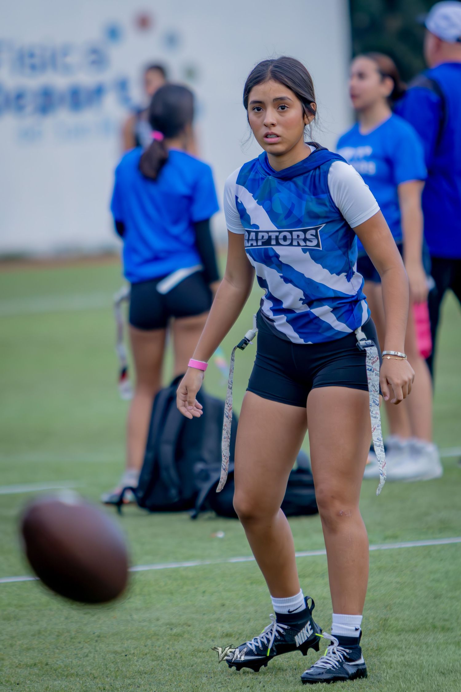 Young female flag football player in Raptors jersey watches a football on a green field during practice.