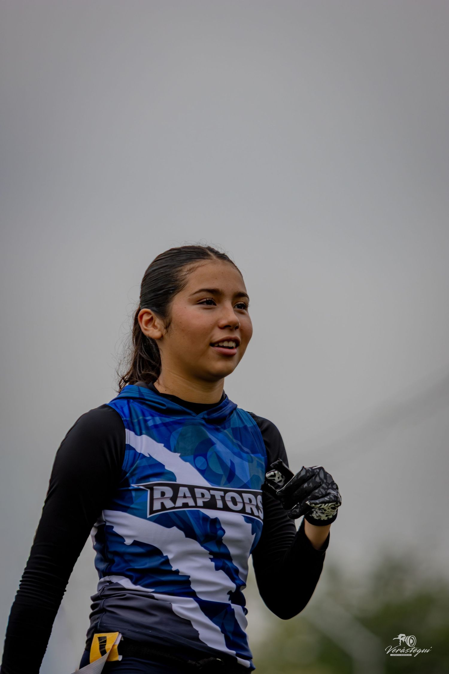 Young female athlete wearing a Raptors blue camo jersey and black gloves during an outdoor sports event.