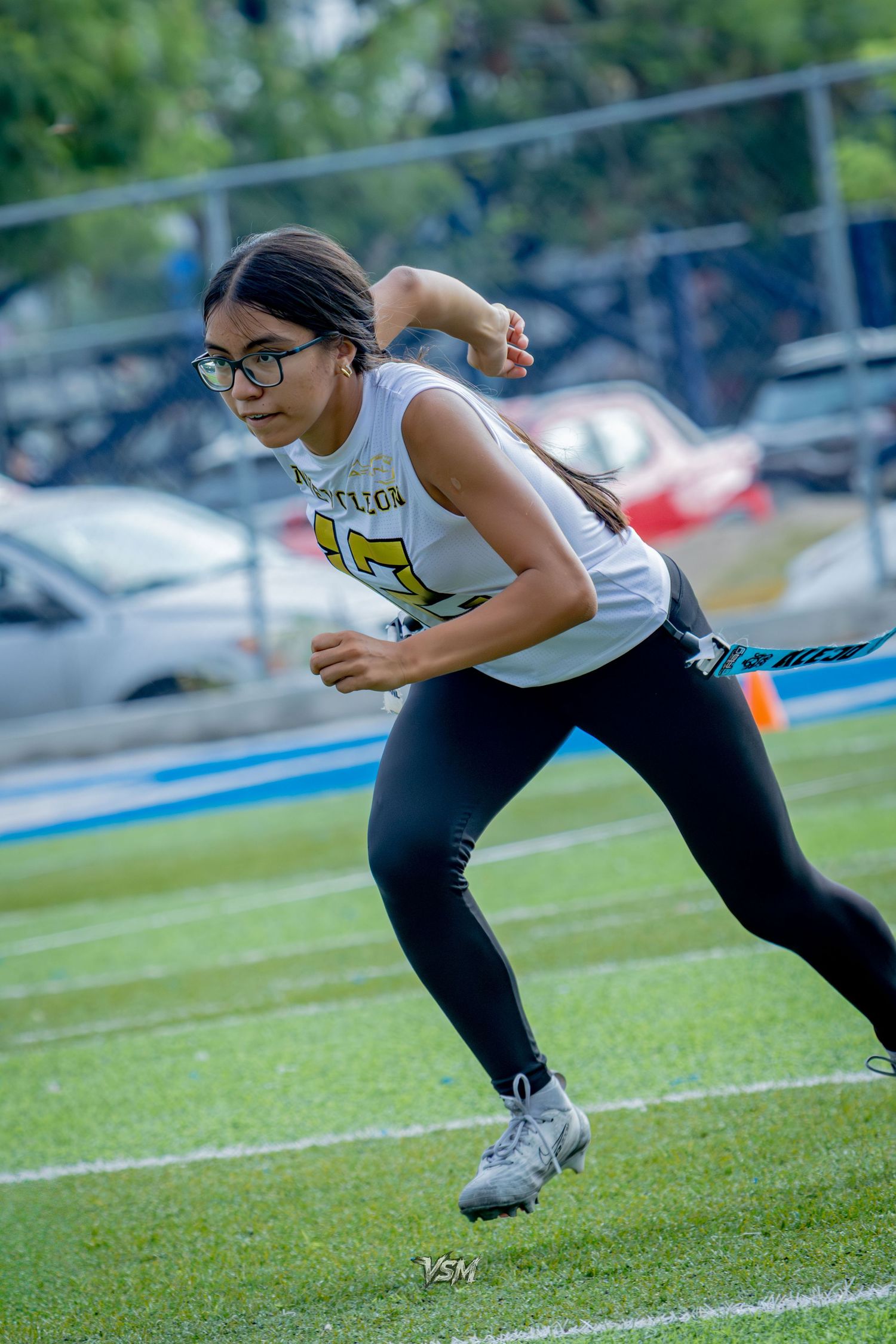Young female athlete in glasses sprinting on green turf field wearing black leggings and white jersey during game.