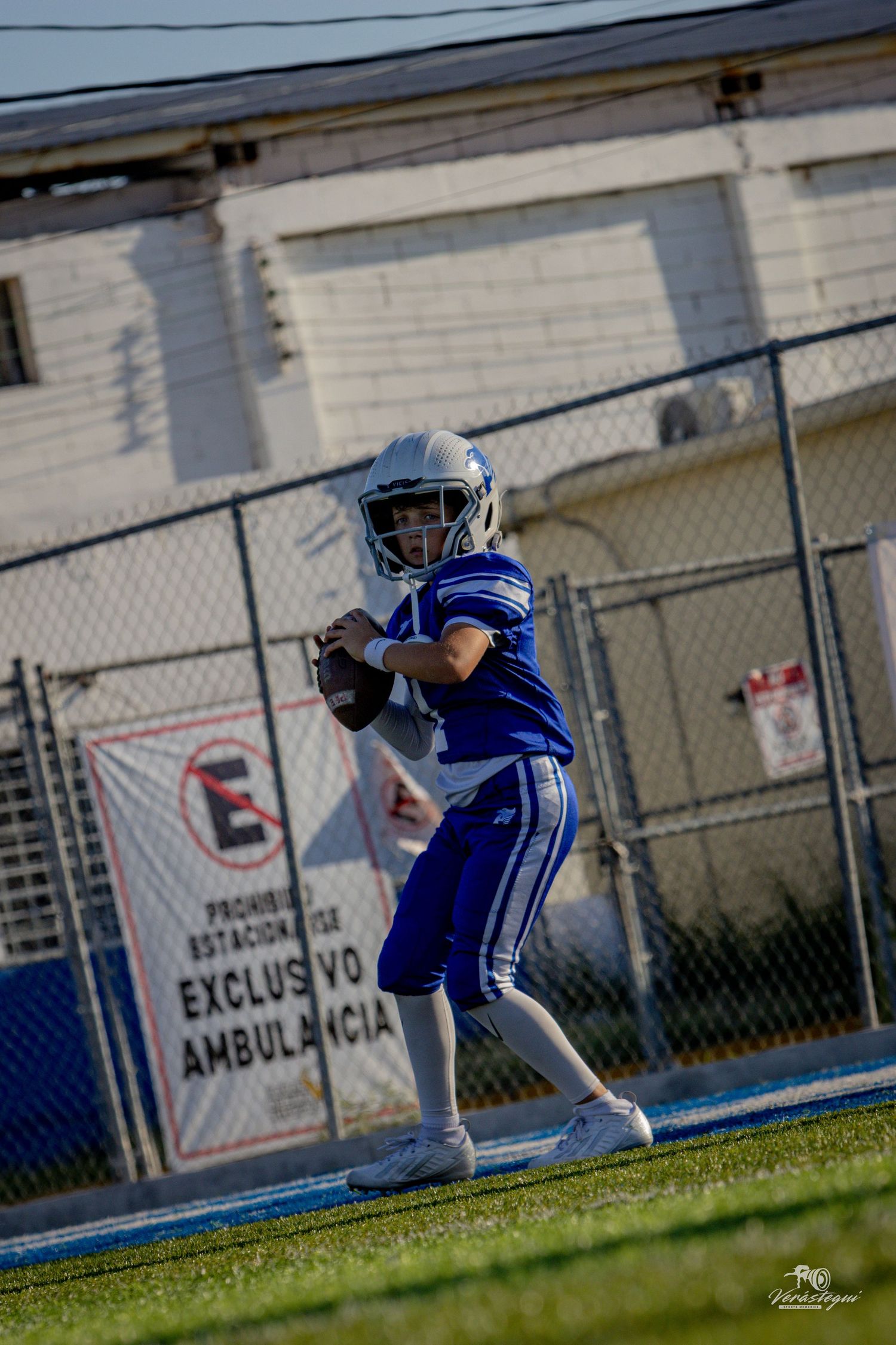 Young quarterback in blue uniform prepares to throw during a youth football game near an ambulance parking sign.