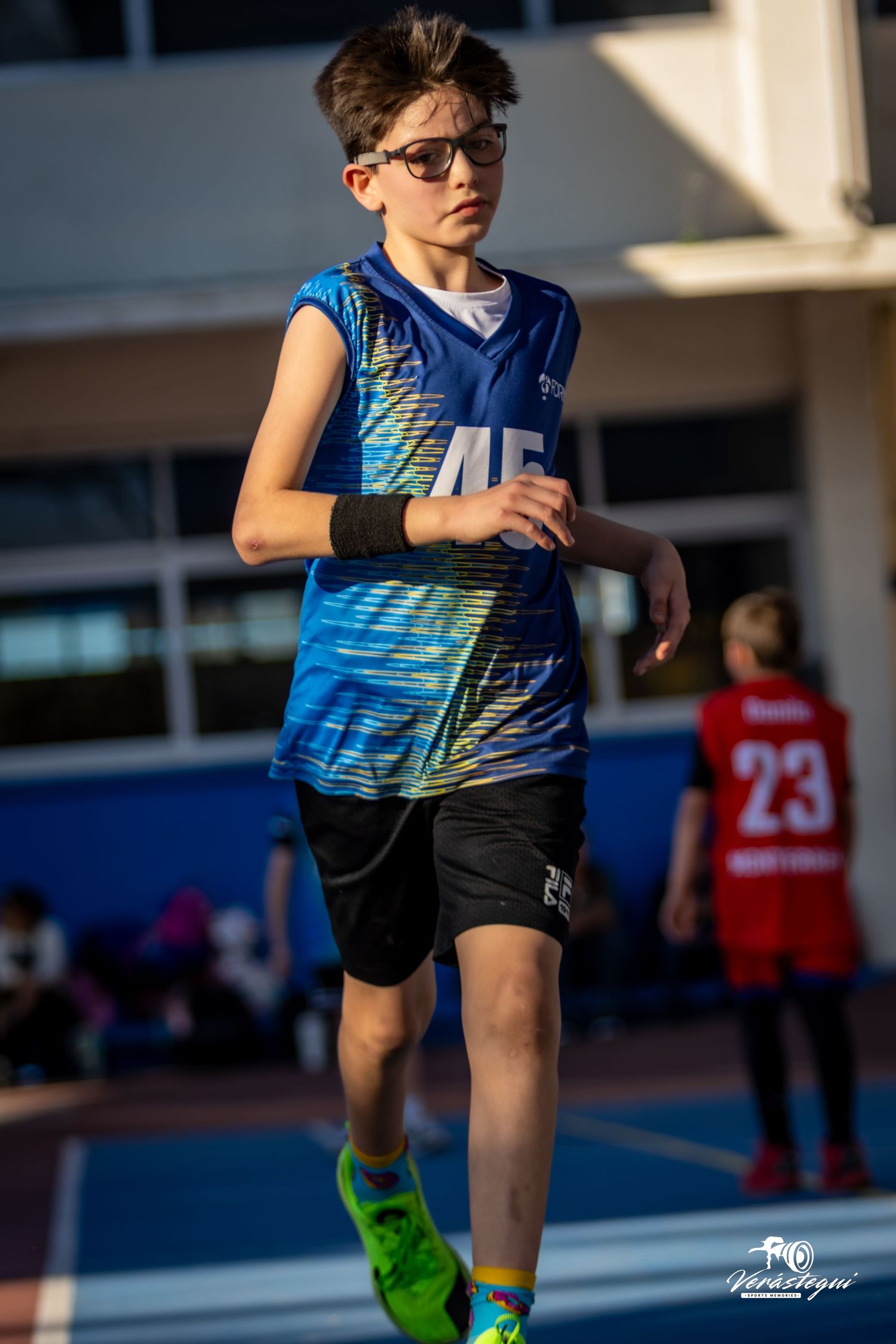 Young athlete in blue basketball jersey and sunglasses runs on outdoor court during daytime competition.