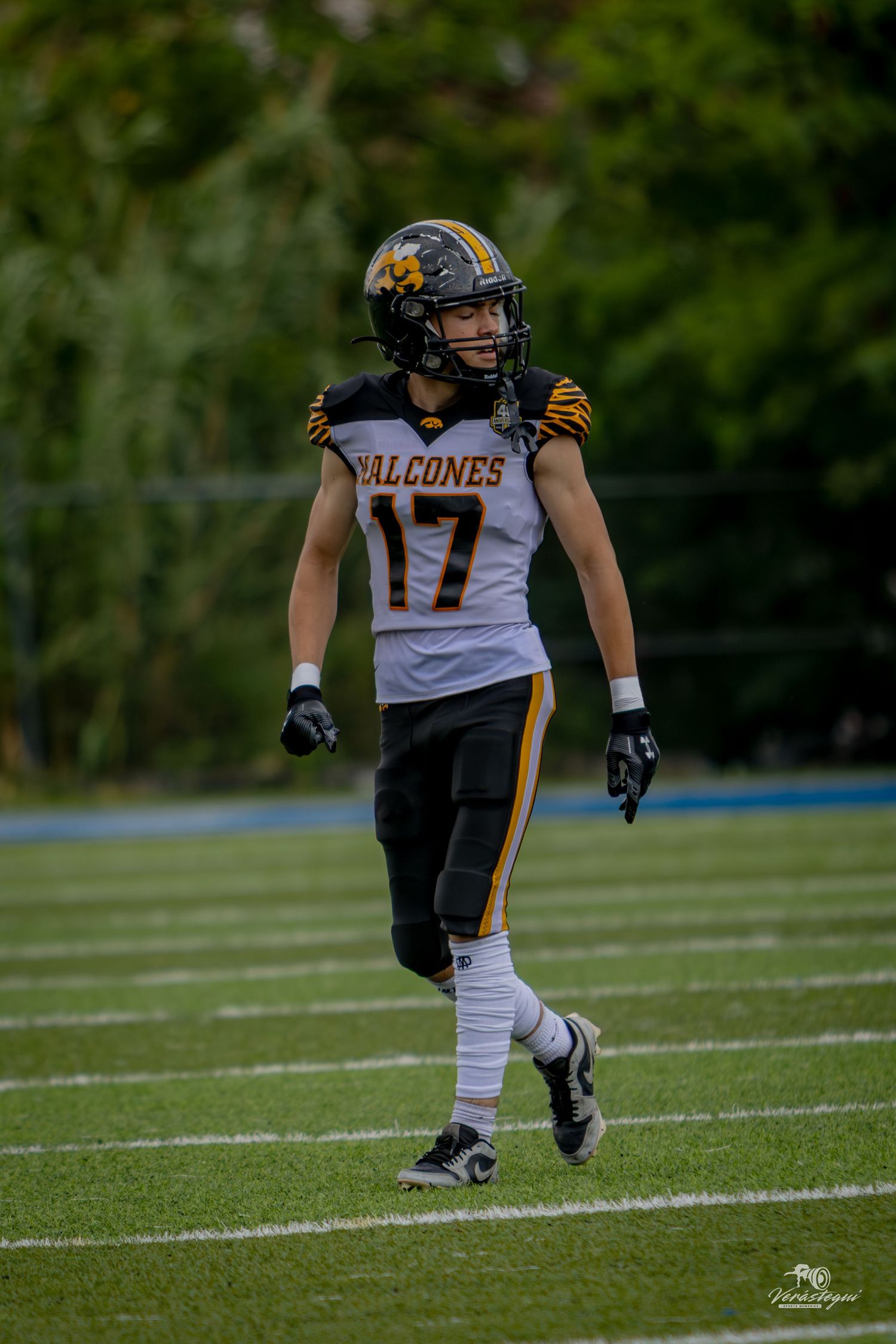 Football player #17 in Falcones uniform walks on field wearing black helmet and gold accents during game.