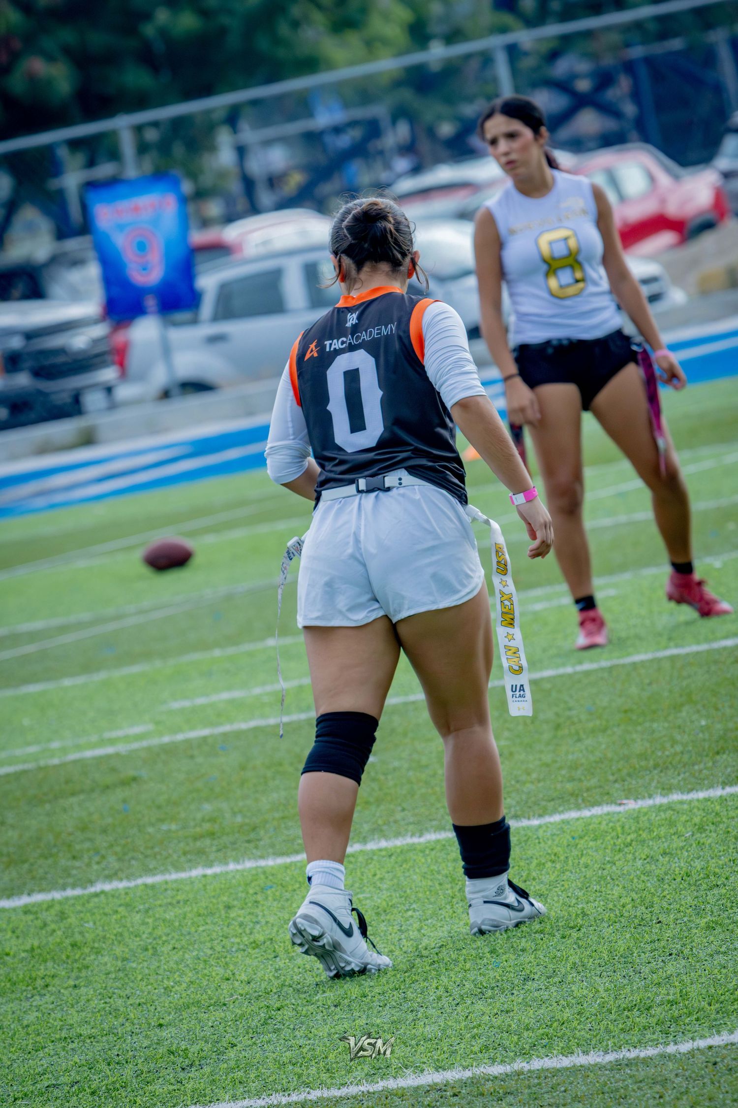 Two female flag football players in jerseys numbered 0 and 8 compete on a green turf field during an outdoor game.