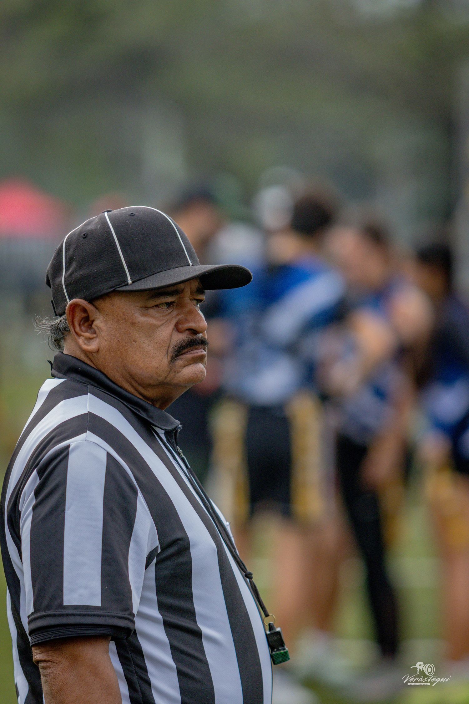A football referee in a black and white striped shirt and cap stands on the field during a game.