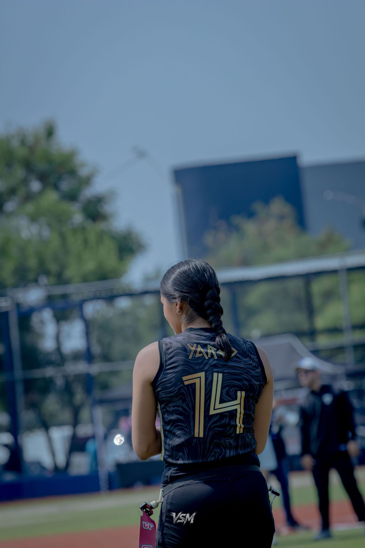 Female softball player #14 wearing black and gold jersey stands on field during outdoor game.