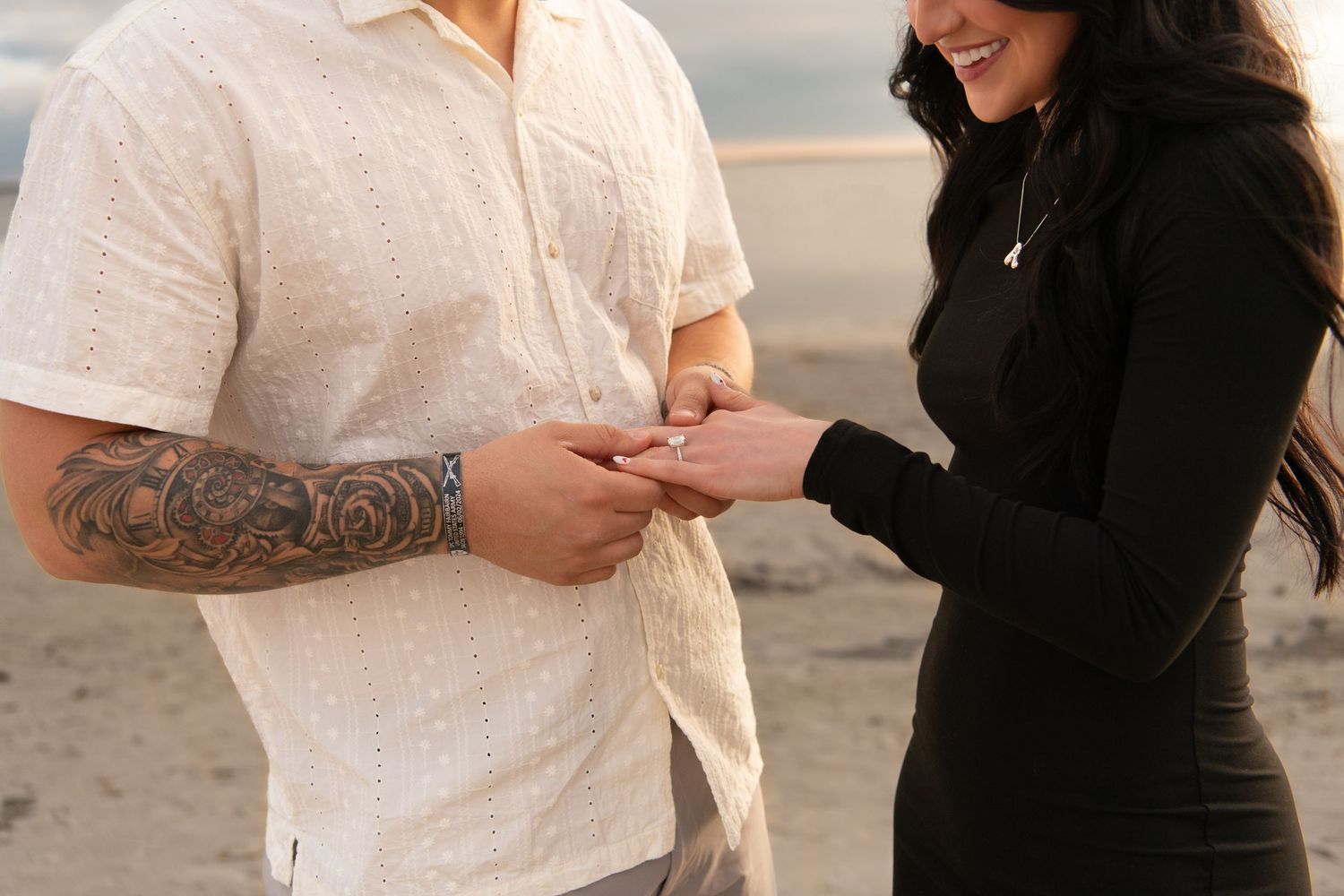 A tattooed man places a ring on a smiling woman's finger on a beach during a romantic proposal.