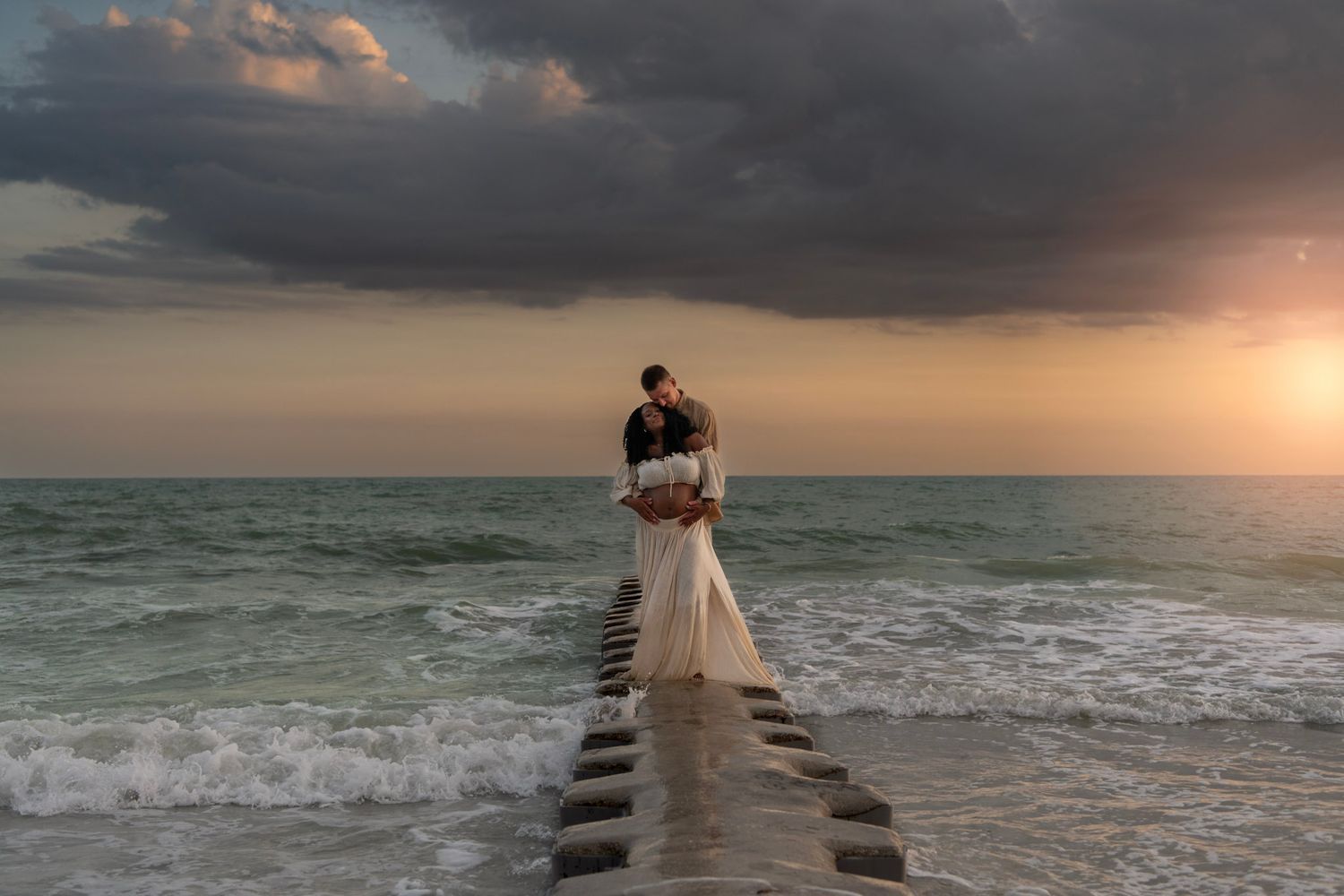 Couple embracing on a seaside pier at sunset, bride in white gown, dramatic stormy sky over ocean waves.