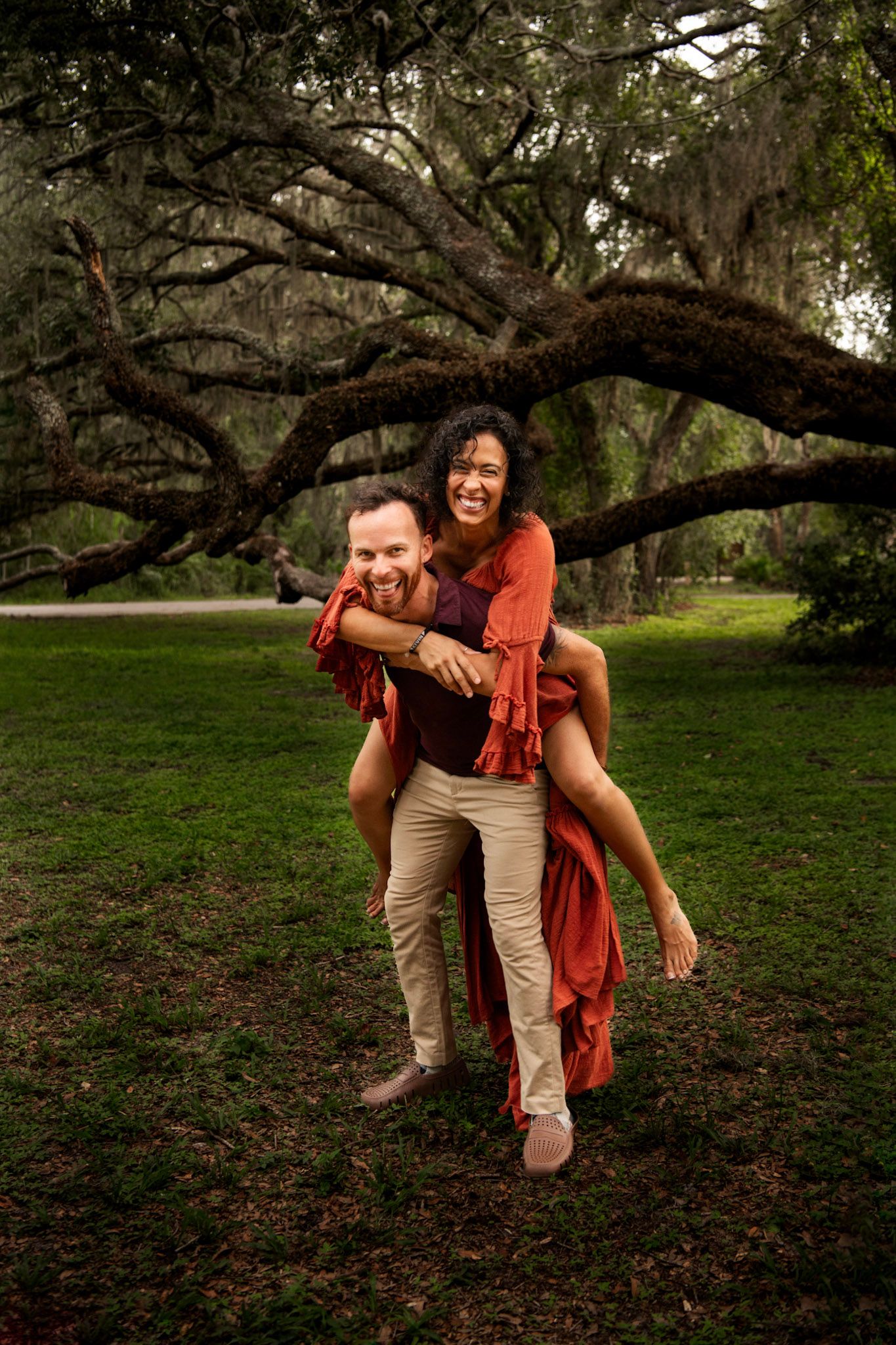 Playful couple portrait beneath sprawling oak trees with Spanish moss in a Florida park setting.