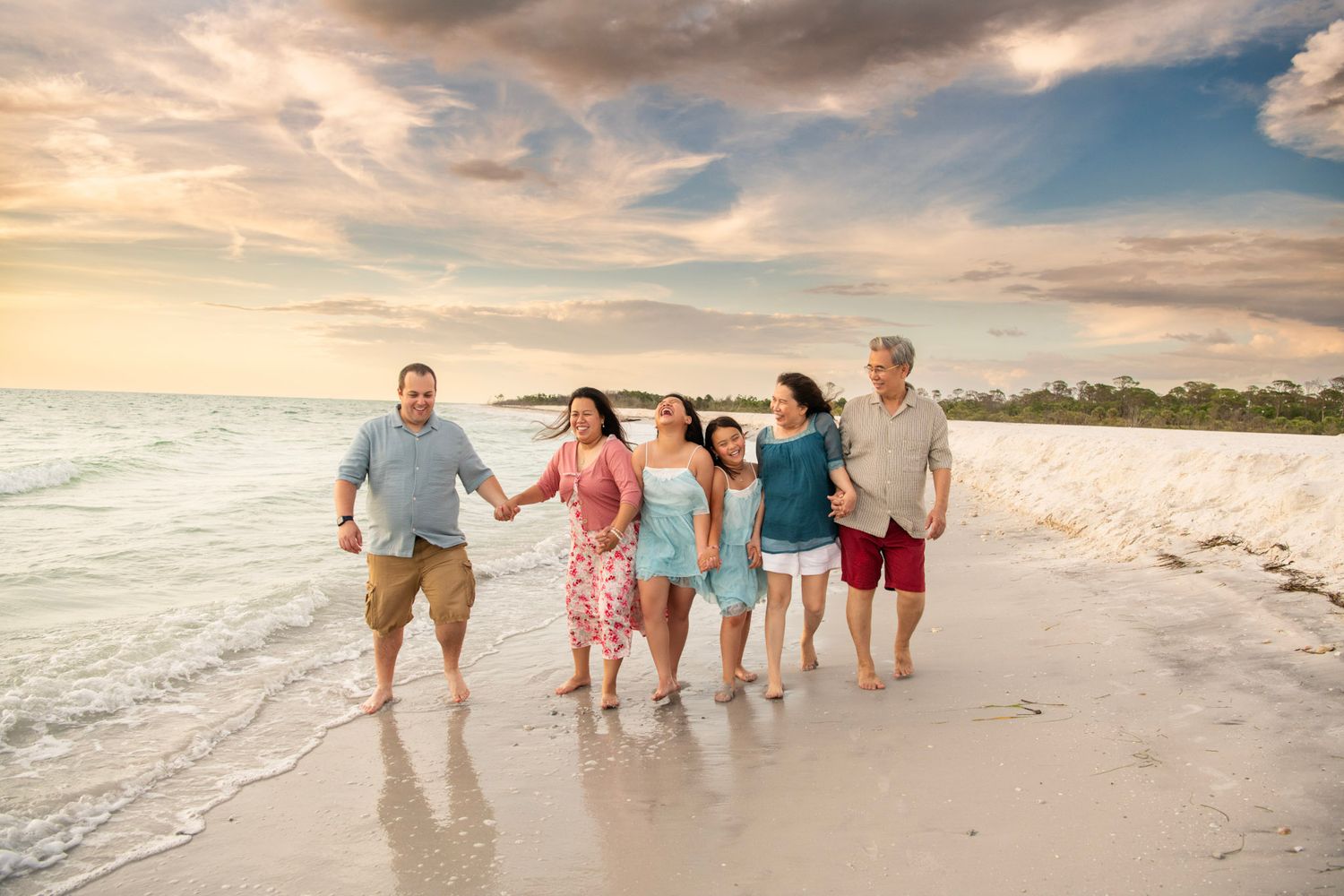 A happy multigenerational family walks hand-in-hand along a sandy beach at sunset with gentle waves nearby.
