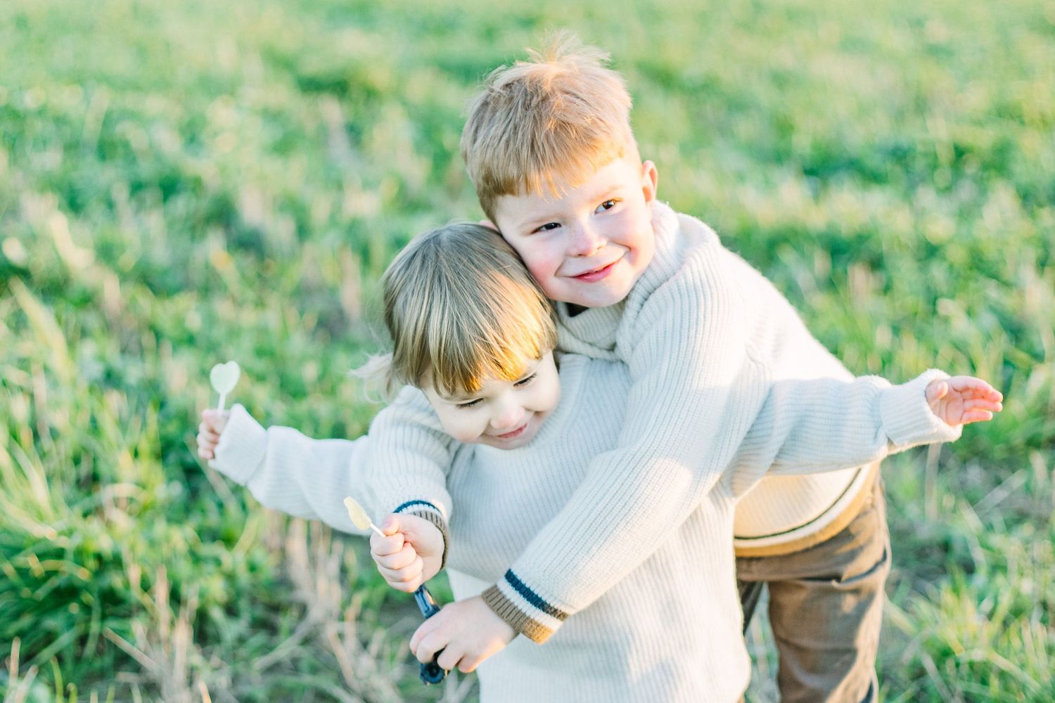 Two children in light colored sweaters play together in a sunny green field while spreading their arms.