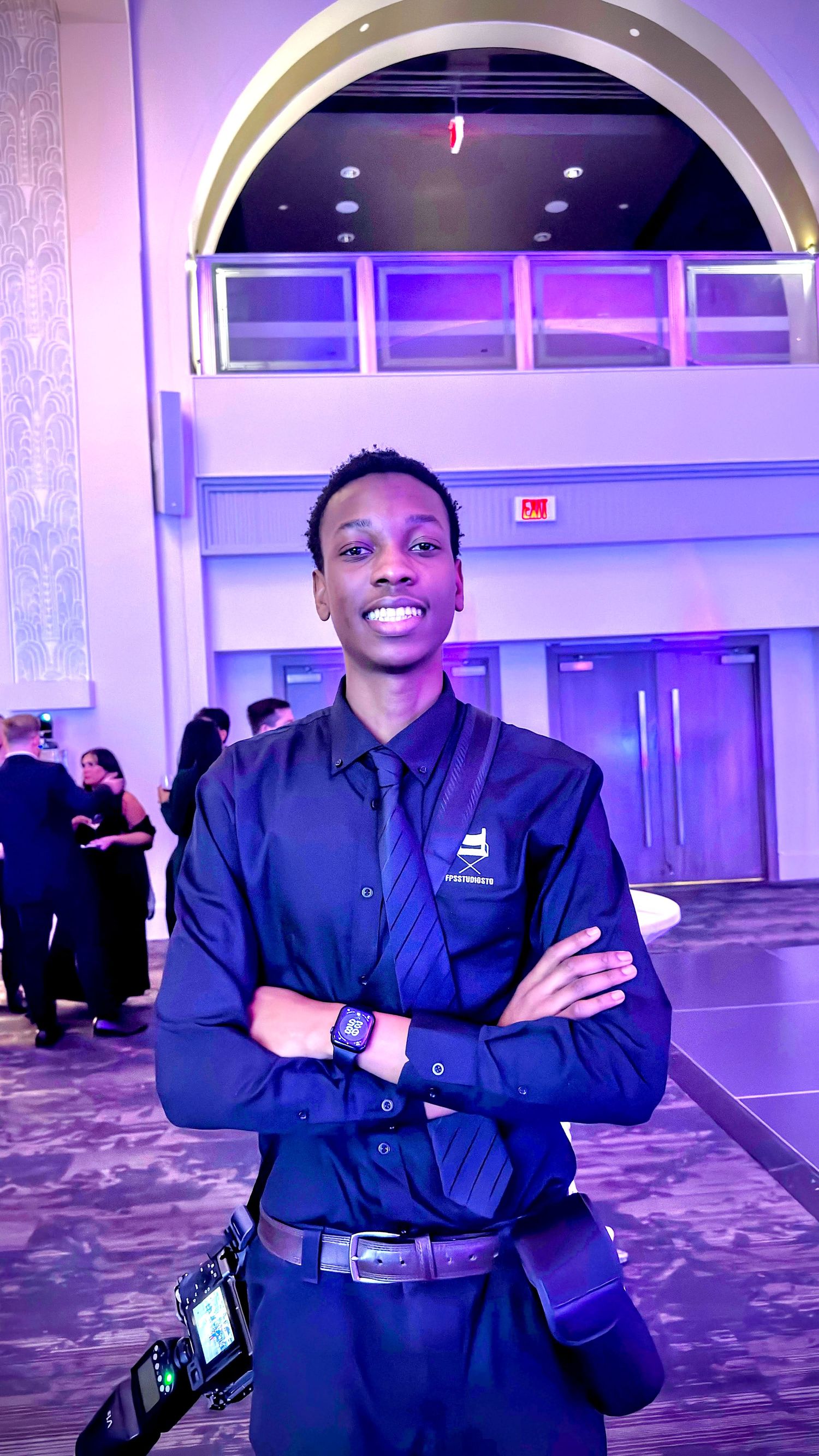 Smiling young Black man in black uniform with tie standing in a purple-lit event hall.