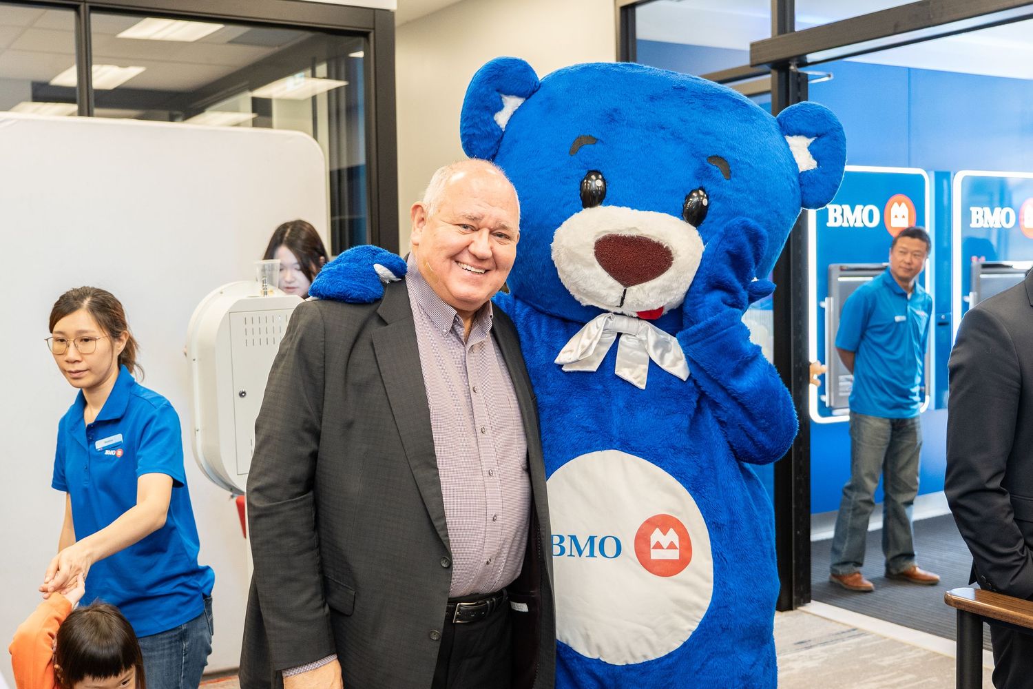Senior man hugging BMO blue bear mascot inside BMO bank branch.