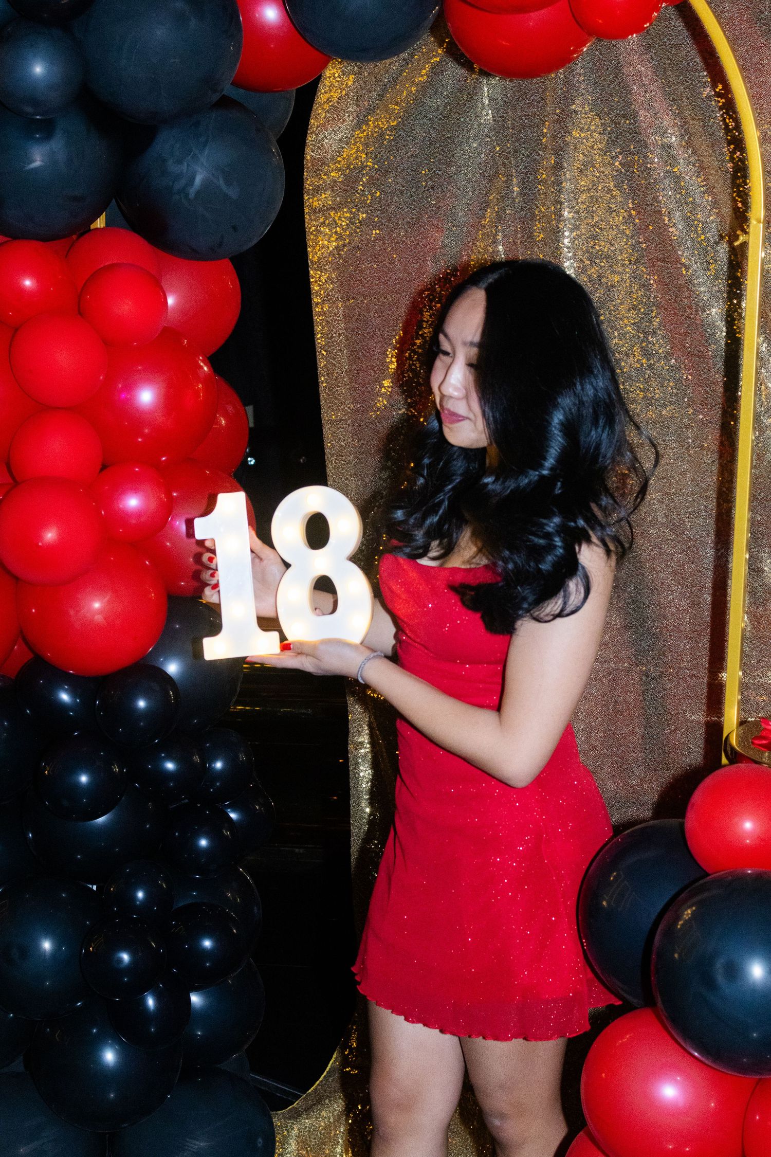 Young woman in a red dress holding illuminated number 18 sign surrounded by red and black balloons.