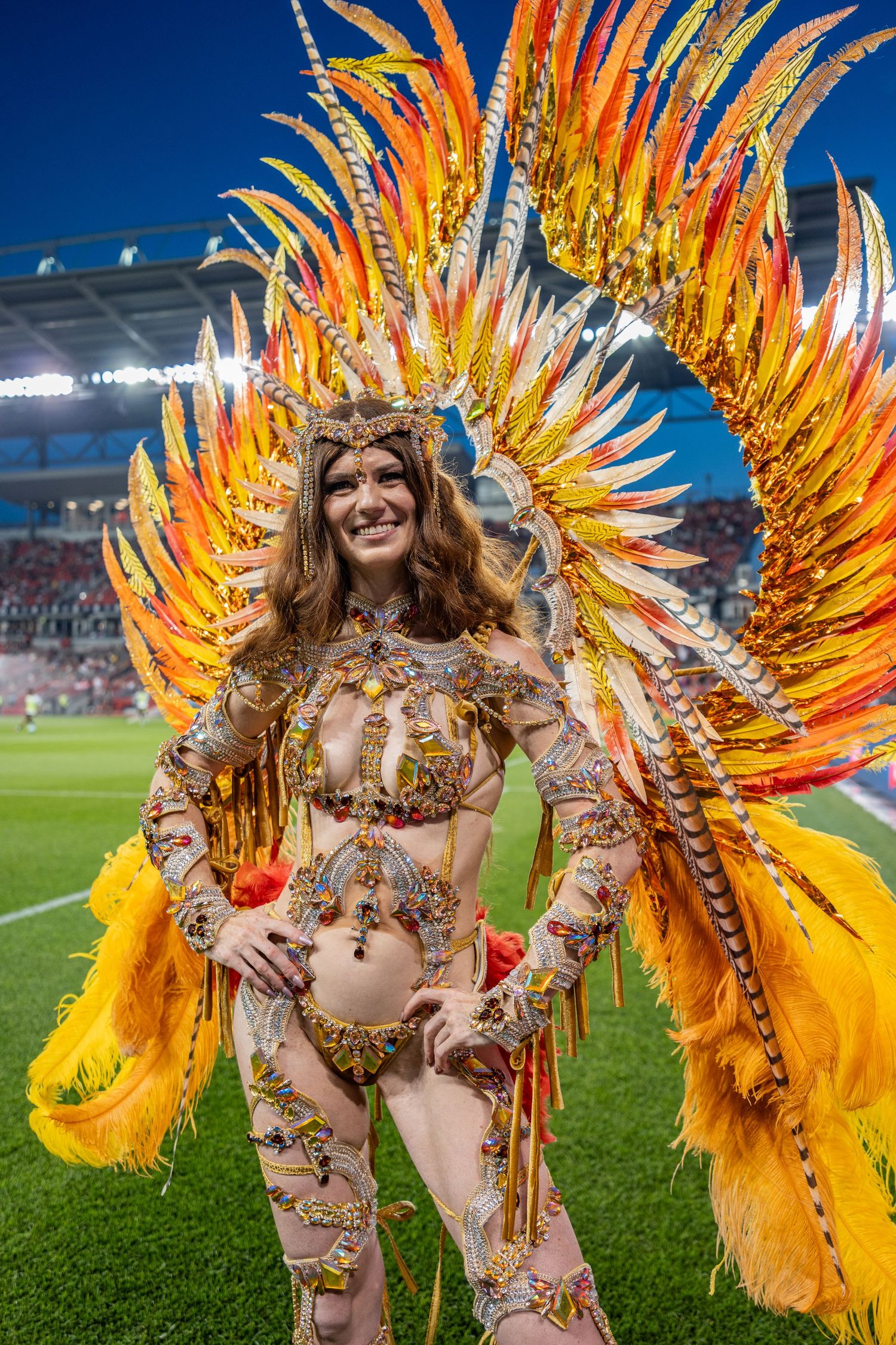 Woman in elaborate golden orange feathered carnival costume posing on stadium field at night.