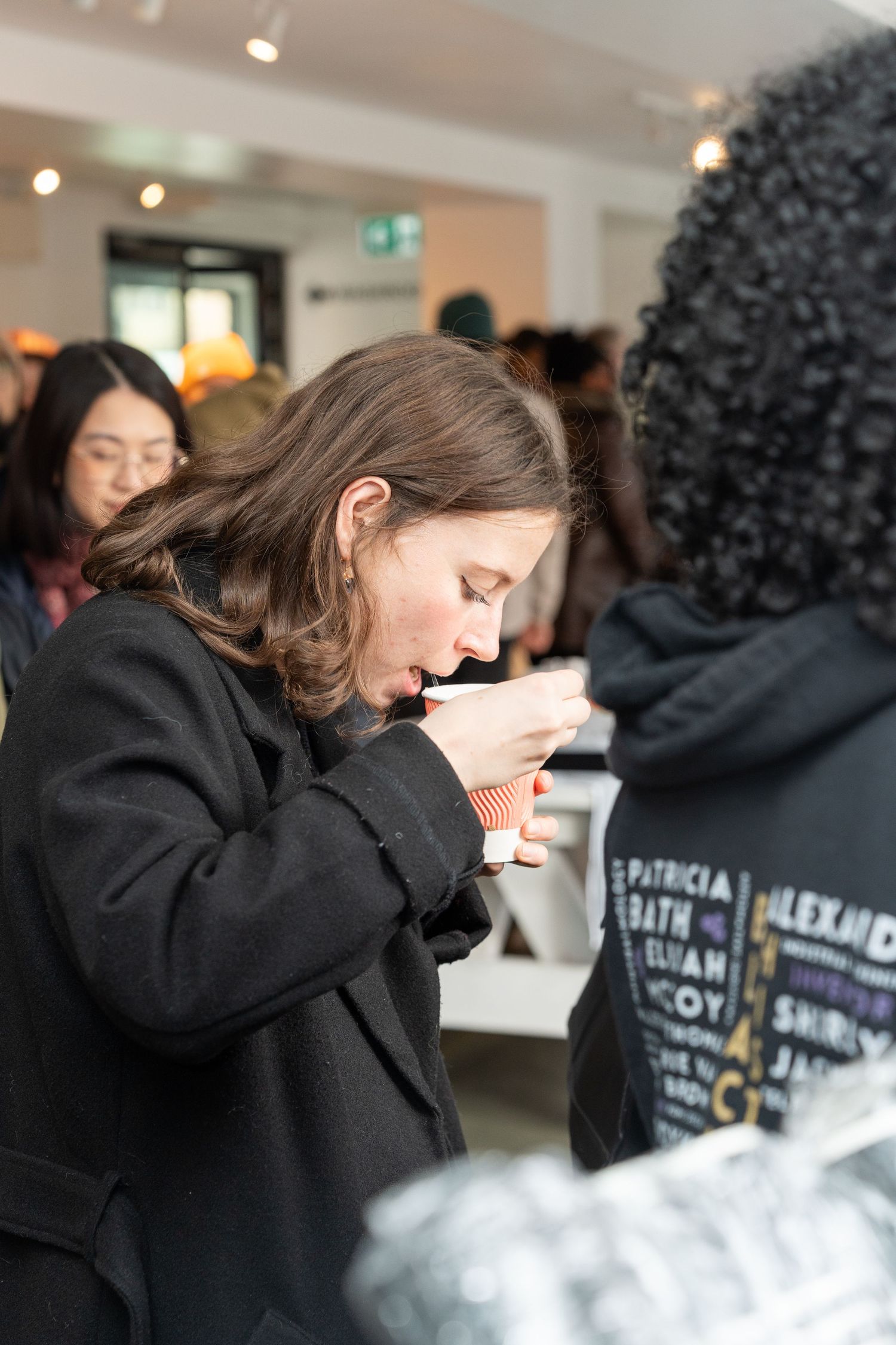 Young woman in black coat examining a small item at a busy indoor vendor market event.