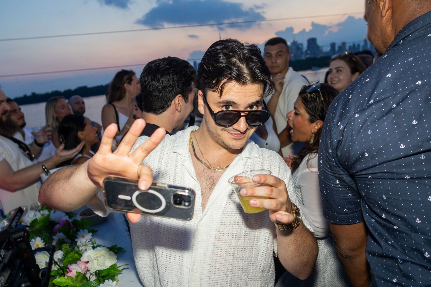 Young man taking a selfie while holding a drink at a sunset rooftop party.