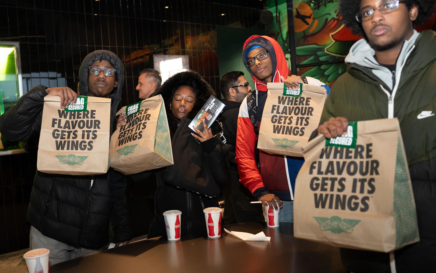 Group of young people holding branded takeout bags reading Where Flavour Gets Its Wings at restaurant event.
