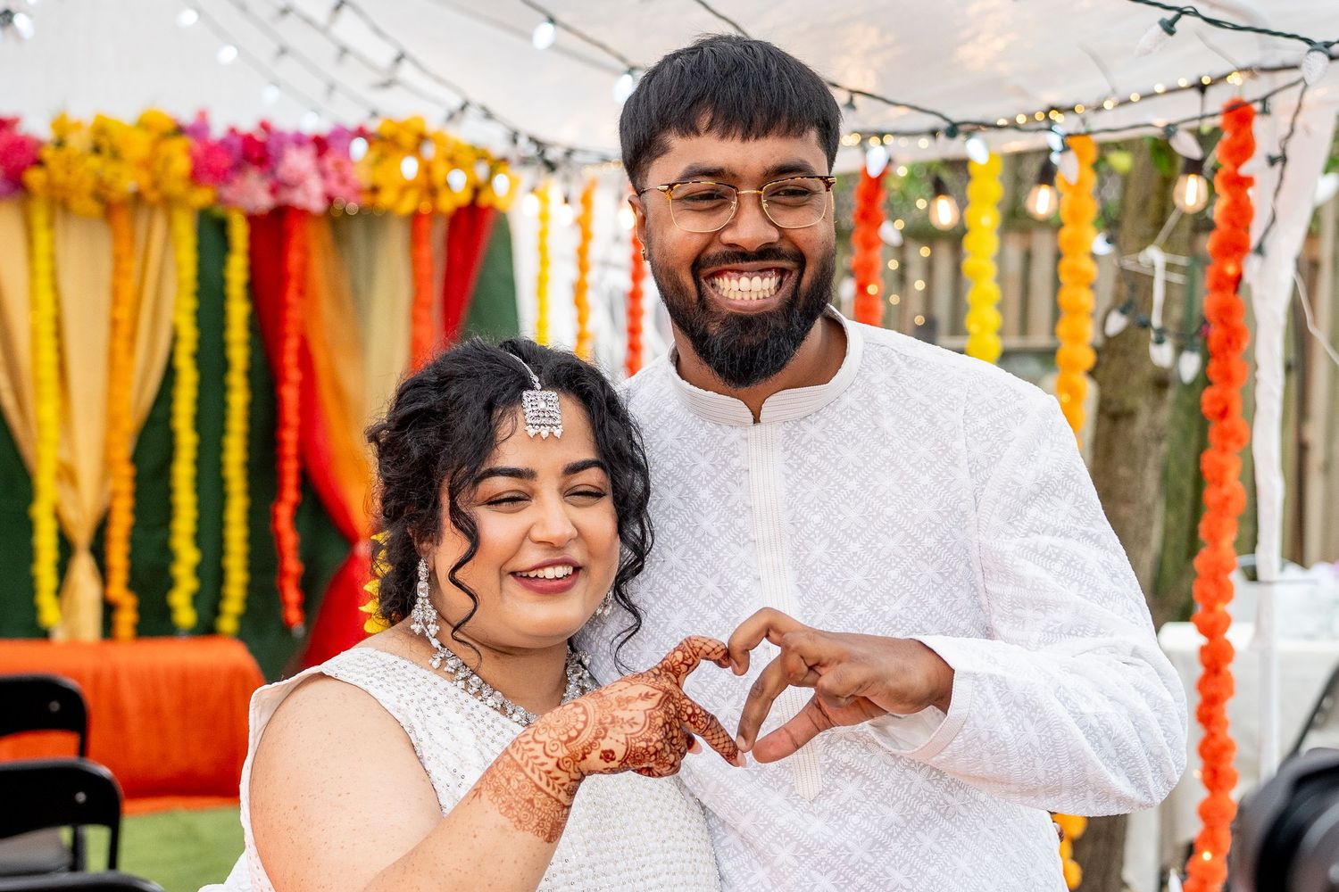 Smiling couple in white outfits making a heart shape with hands at Indian wedding celebration.