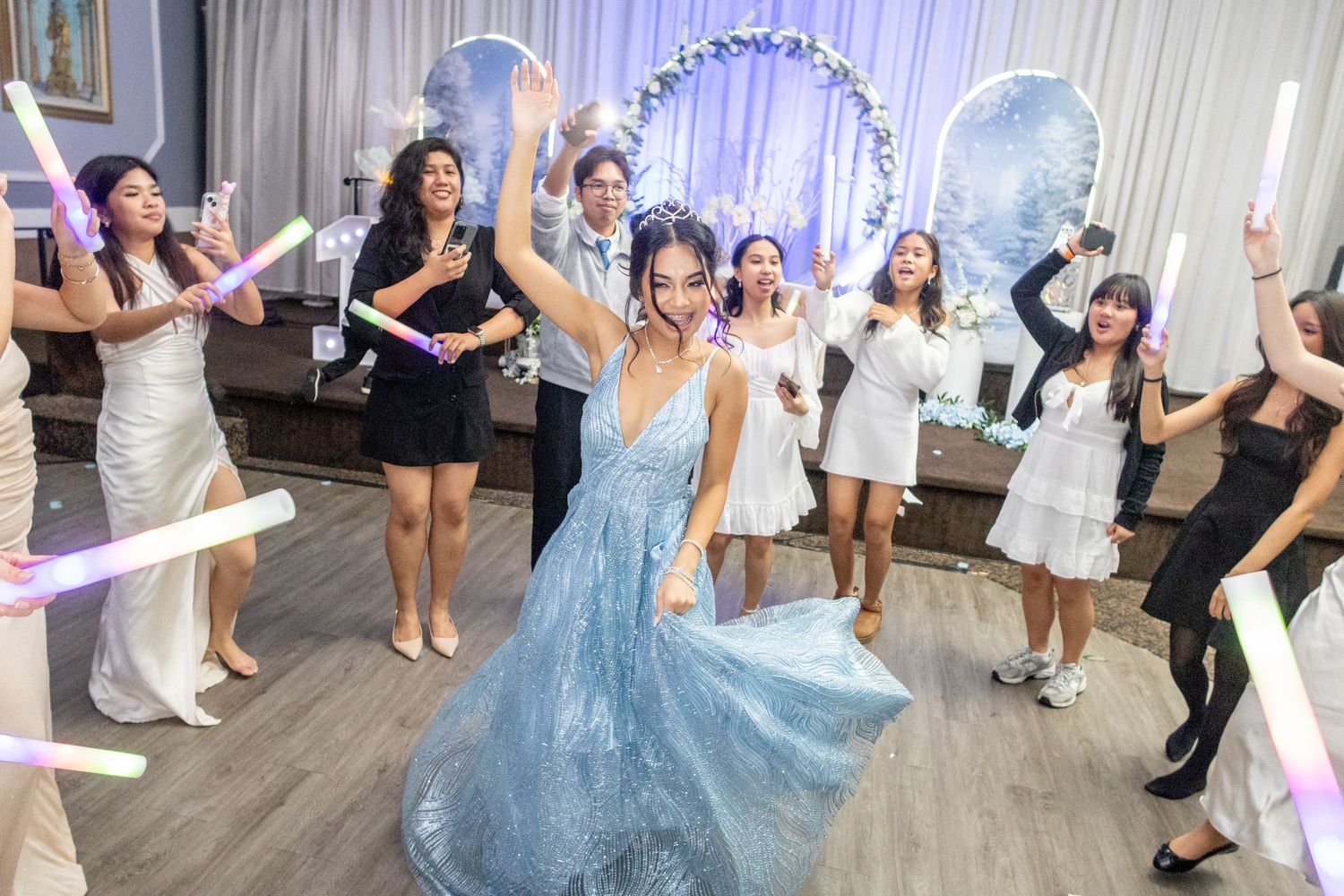 Young woman in blue gown dancing joyfully with friends holding glow sticks at a festive celebration.