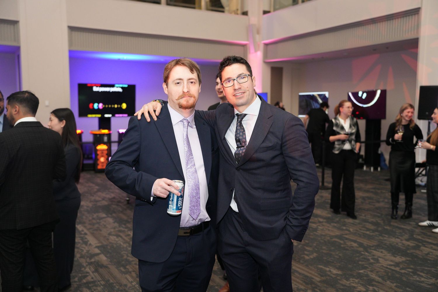 Two men in suits smiling together at an upscale evening networking event with arcade games in background.