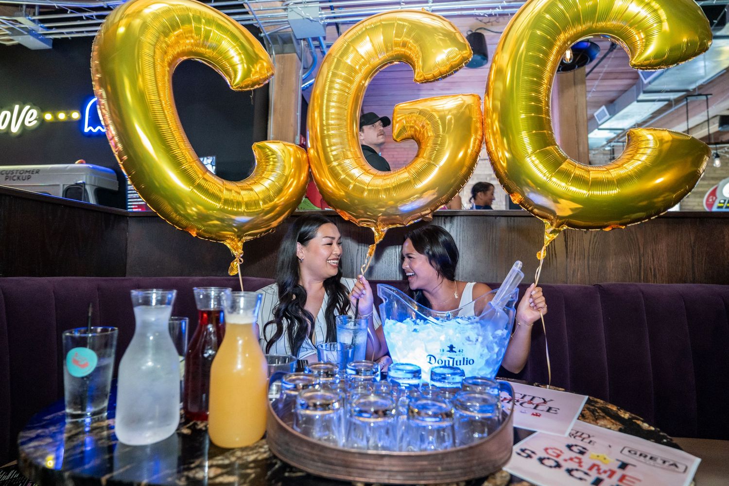 Two women celebrating at bar booth with gold CGC letter balloons and drinks on table.
