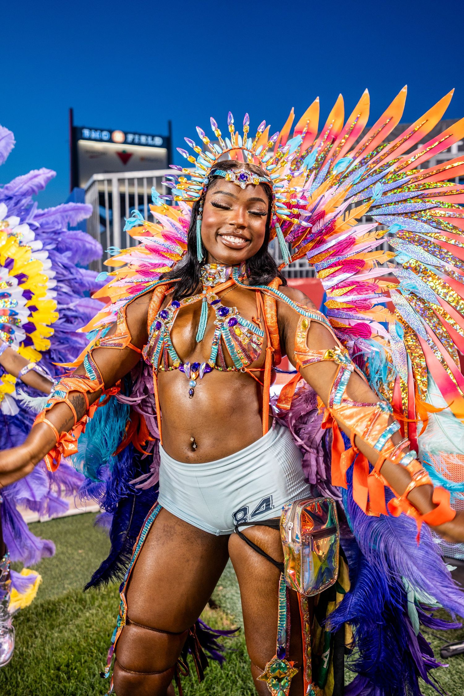 Smiling woman in colorful orange and purple feathered carnival costume at BMO Field festival.