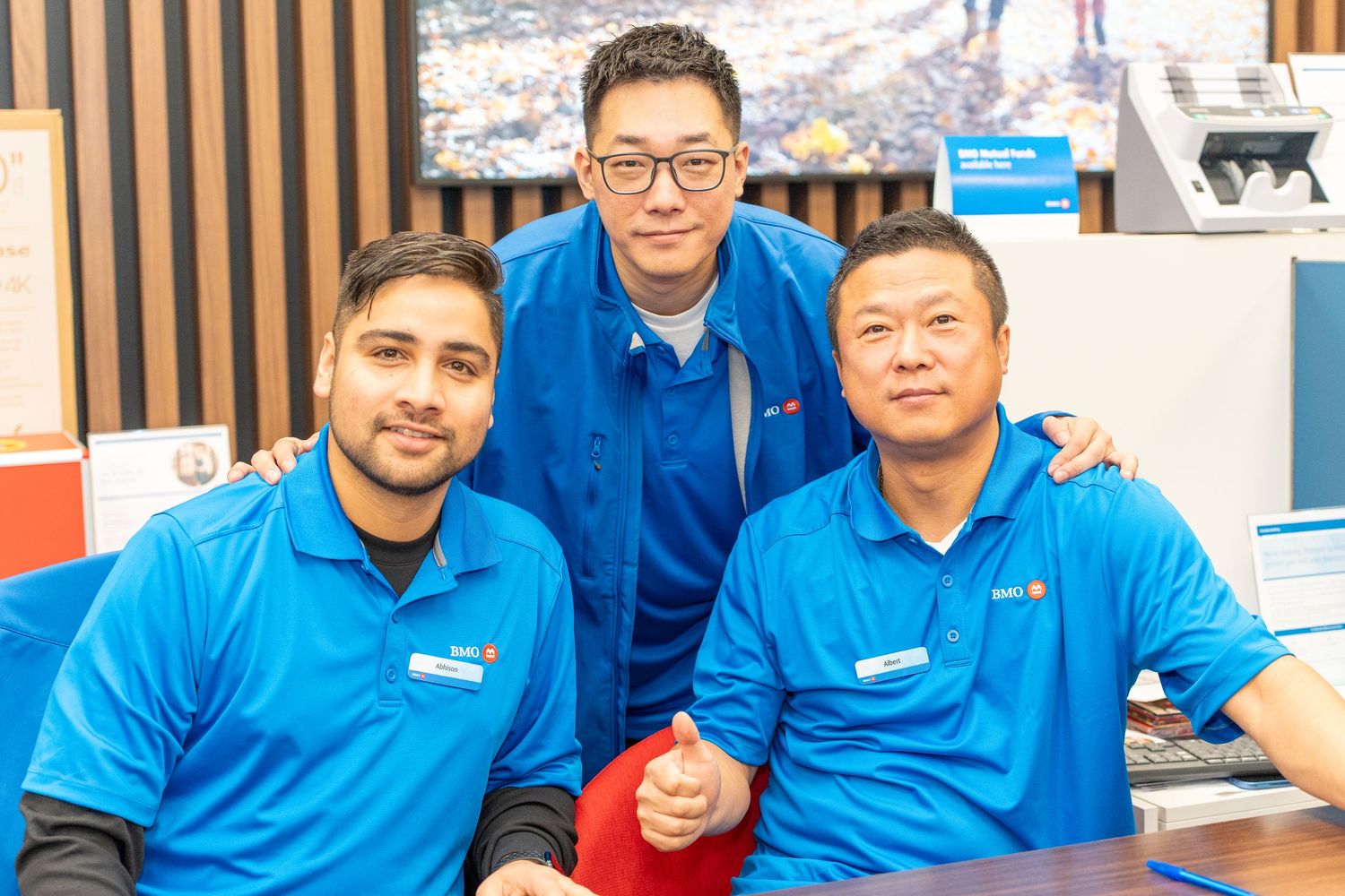 Three BMO bank employees in blue uniforms smile together at a bank branch counter.
