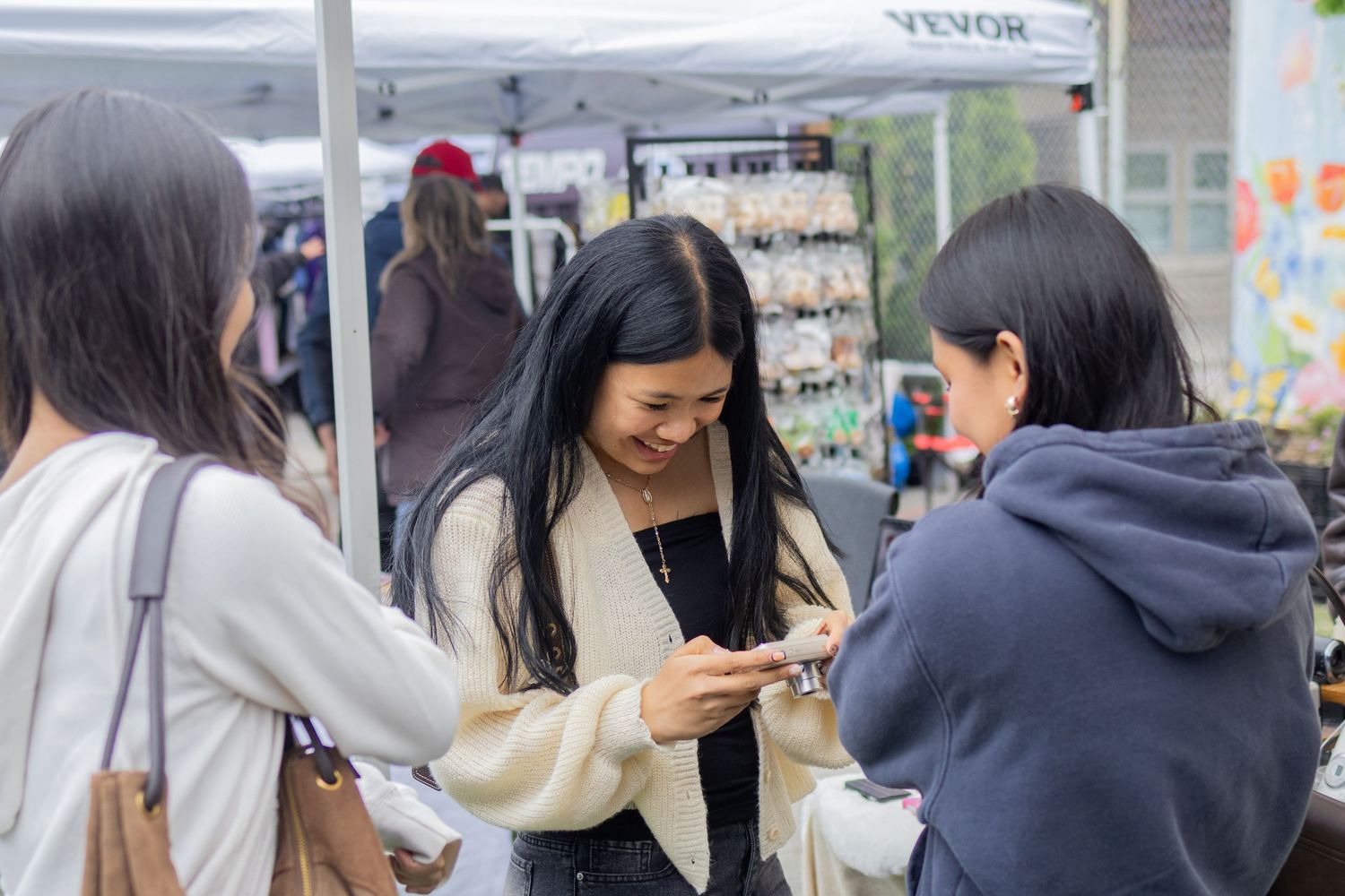 Women examining small items on a smartphone at a busy outdoor market stall.