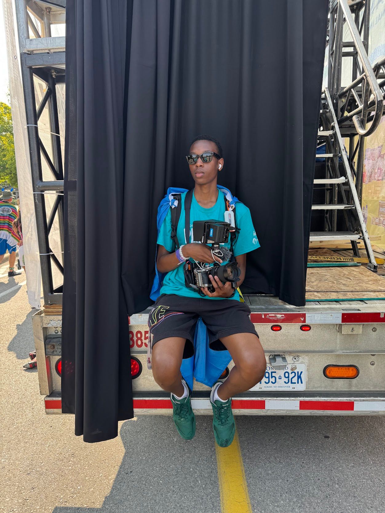 Young Black person in teal shirt sitting on truck bed holding camera at outdoor festival.