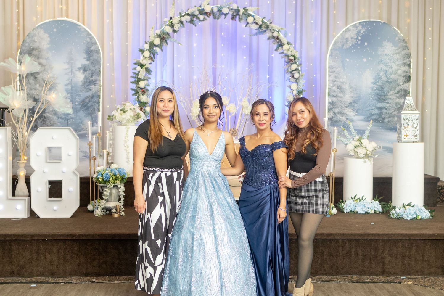 Four women posing together in formal gowns in front of a decorated backdrop at a quinceañera celebration.