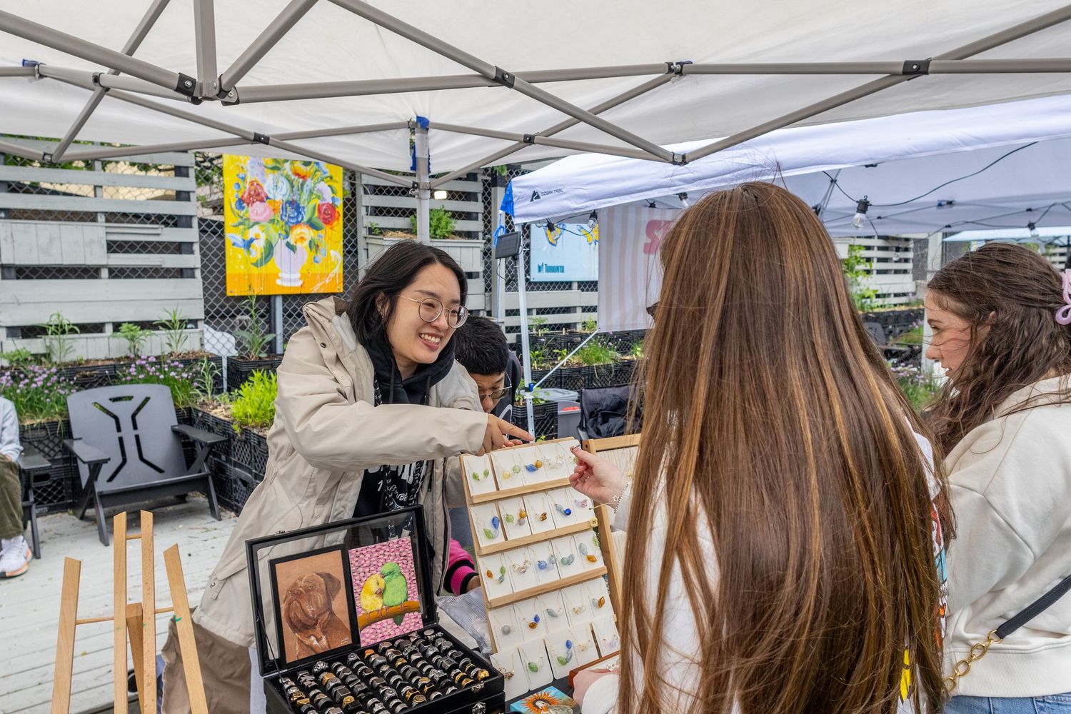 Vendor showing jewelry and artwork to customers at an outdoor market under a white canopy tent.