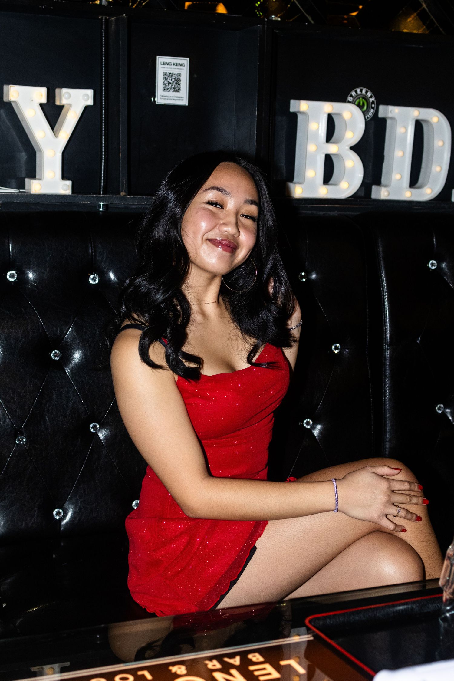 Young woman in red sparkly mini dress smiling at birthday party, seated on black tufted leather couch.