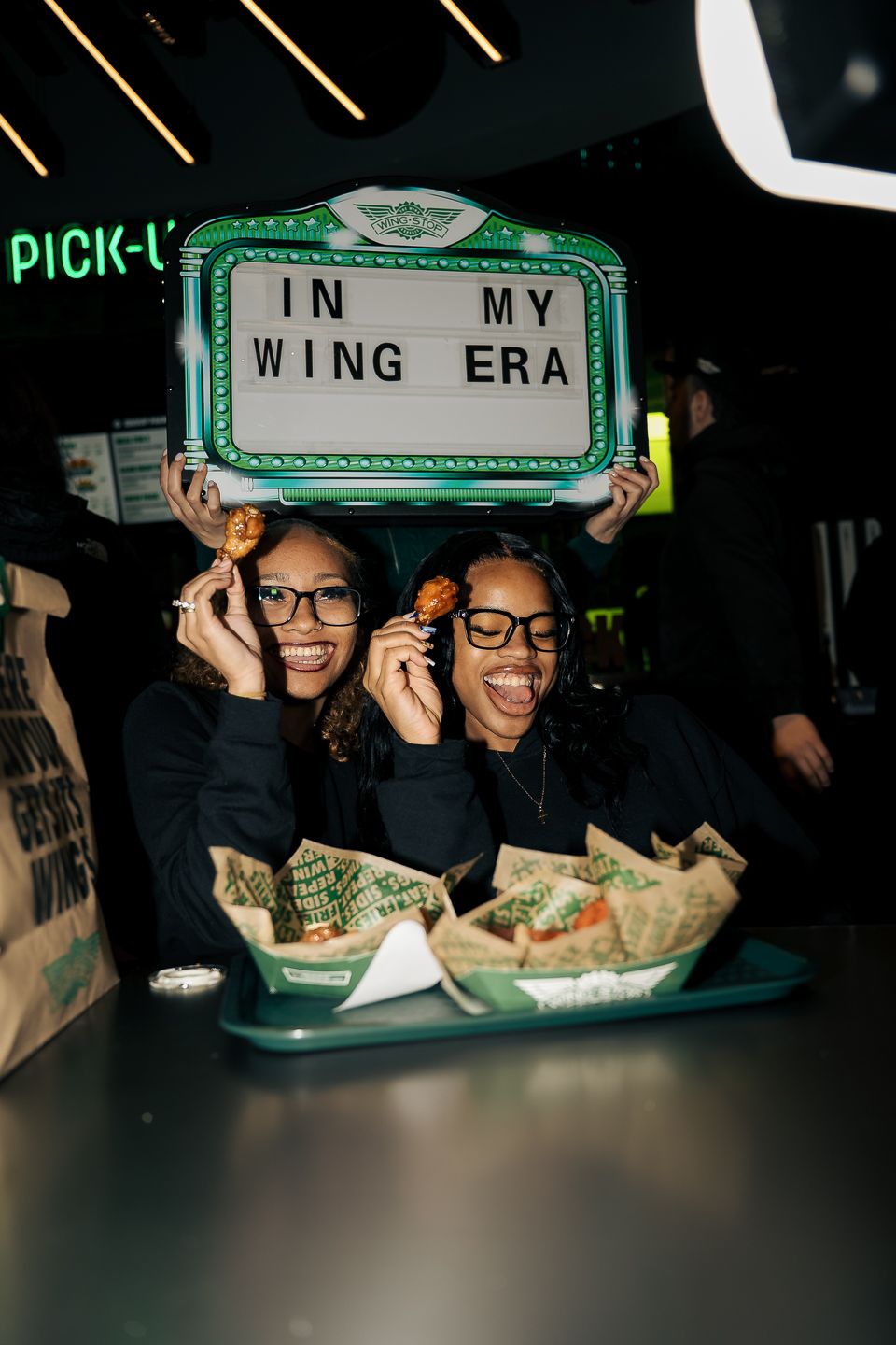 Two women holding 'In My Wing Era' sign while showing chicken wings at Wings restaurant.